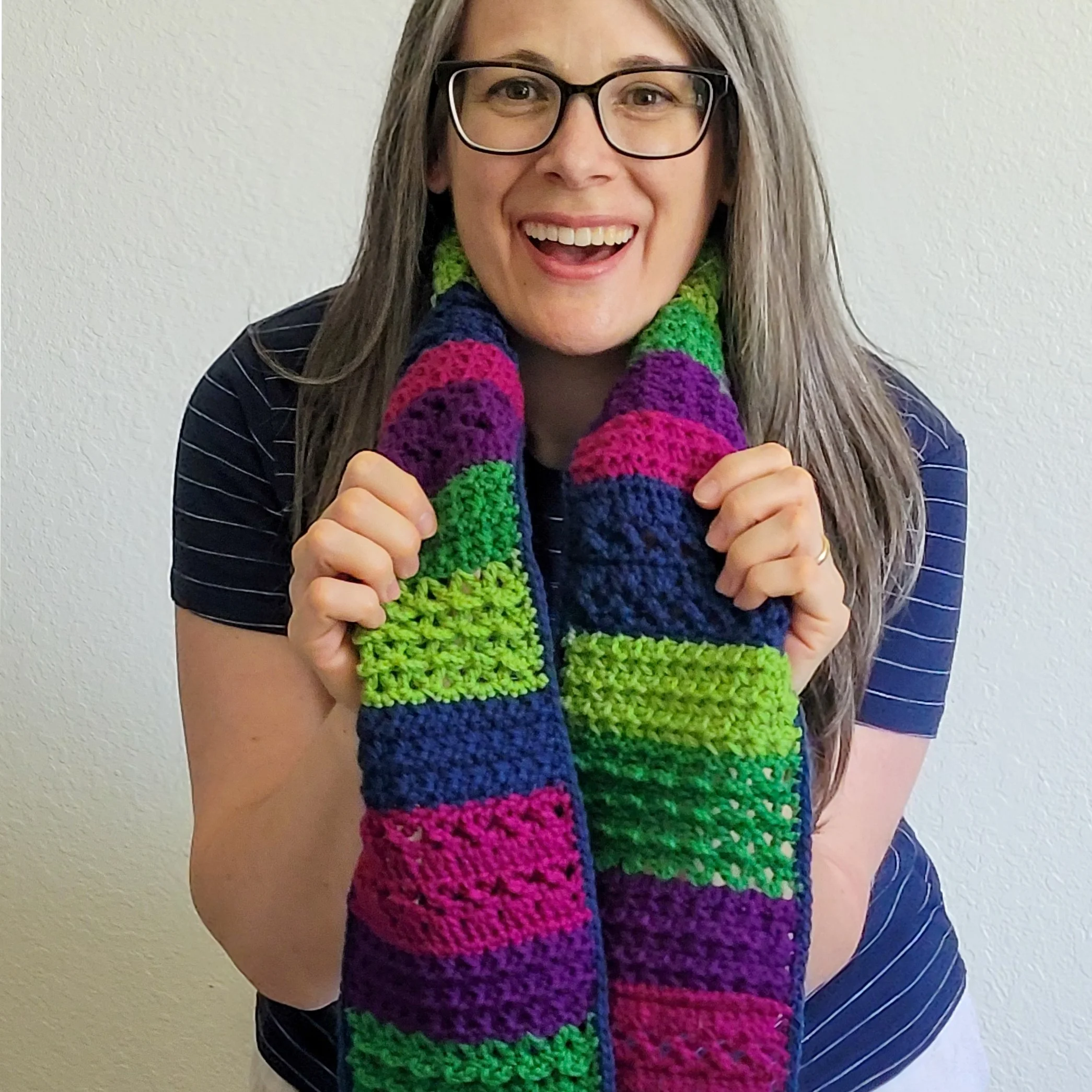 A smiling woman models a colorful crocheted scarf featuring alternating textured stitch patterns in jewel-tone shades of purple, pink, blue, and green. The interplay of color and texture creates a lively, dimensional design.