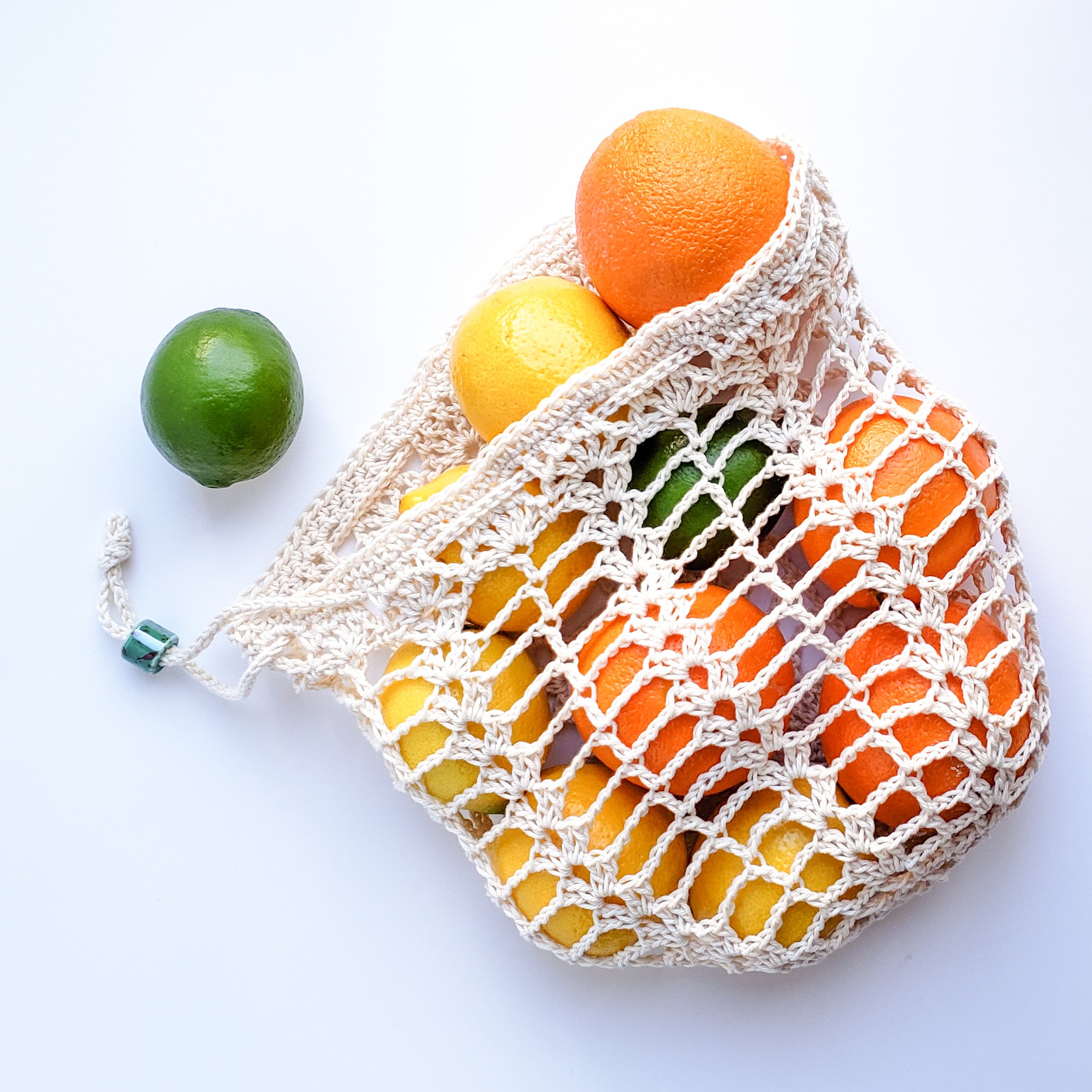 An off-white reusable crochet shopping bag filled with oranges, lemons, and limes. The bag is made with a crochet lace mesh pattern, with a drawstring closure. A single green lime sits to the left of the bag.