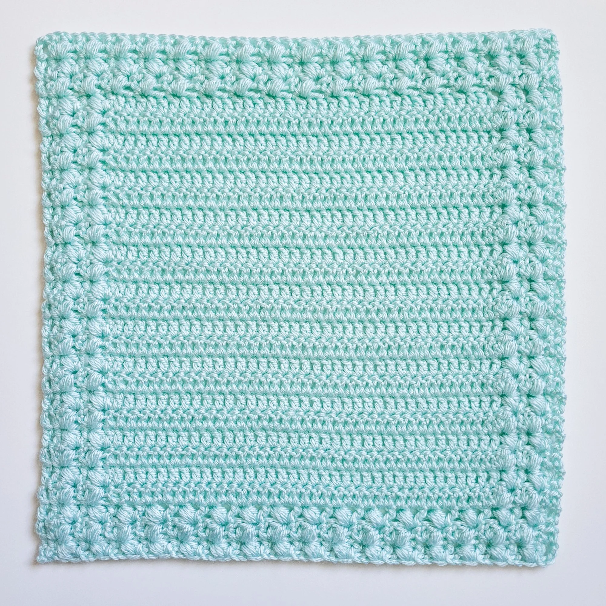 A small, light blue crochet lovey blanket. The design features a V-stitch variation using puff stitches that form a soft, squishy fabric. The textured border is worked seamlessly alongside the center panel as you go.