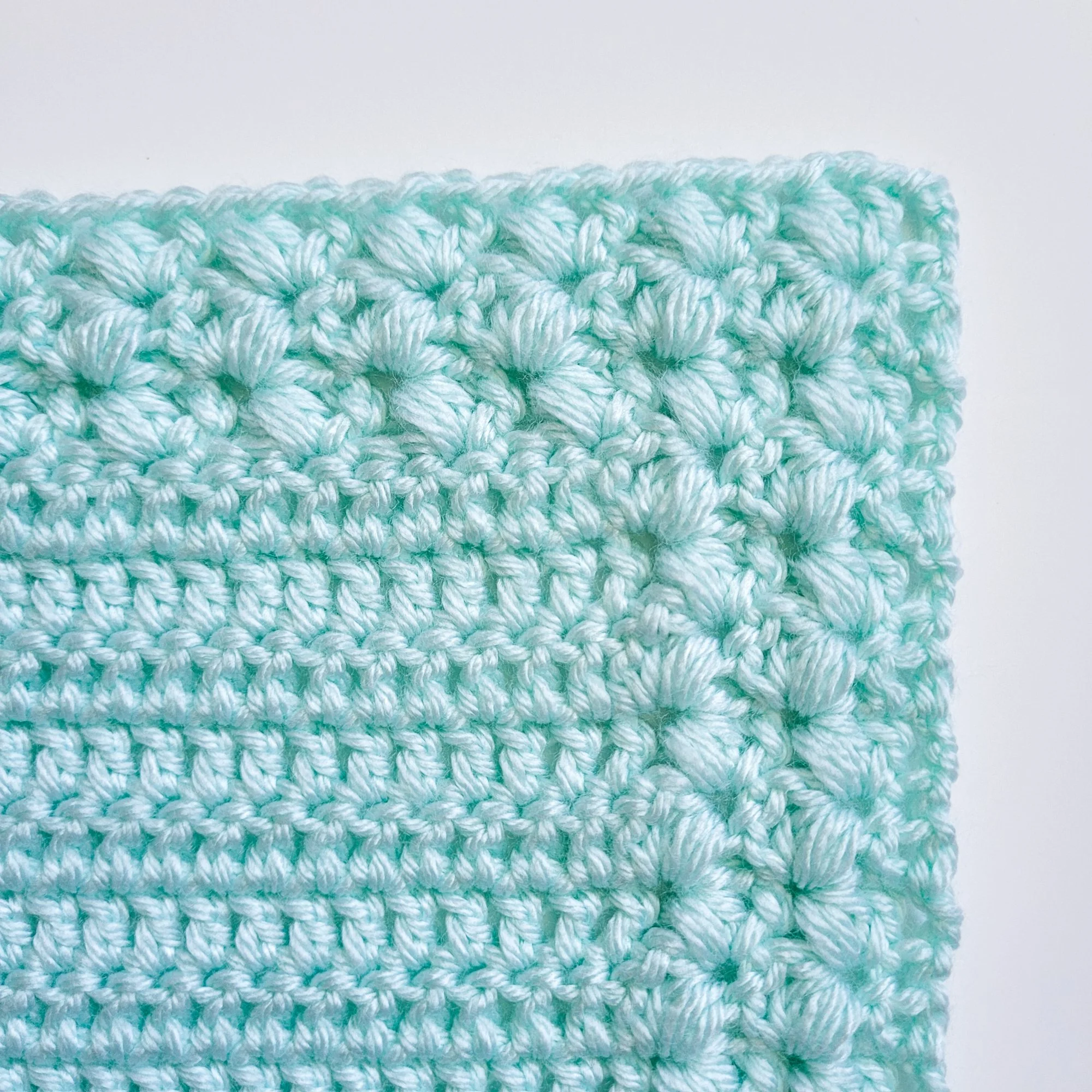 Close-up of the corner of a small, light blue crochet lovey blanket. The textured border, worked seamlessly alongside the center panel, frames the piece with soft, plush detail.