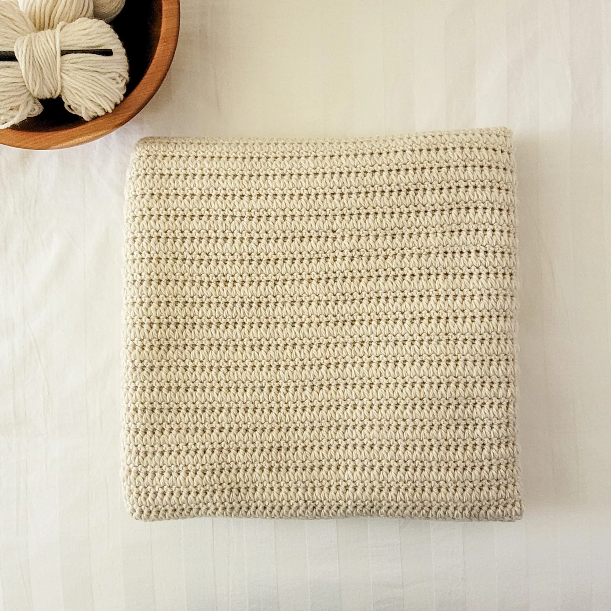 A light beige crochet blanket is folded neatly on a white comforter. The blanket has a distinct ridged texture.