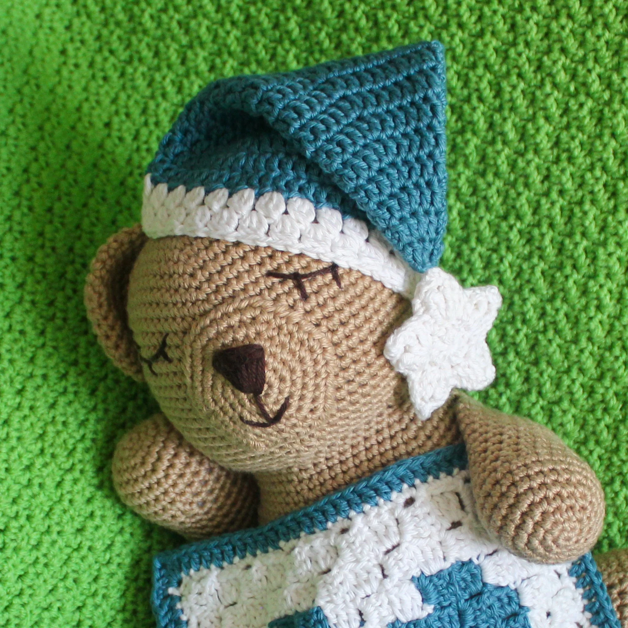A tan crochet bear, with sleepy eyes, nose, and mouth embroidered in black, reclines with his head resting on one arm atop a bright green crochet blanket. He wears a blue crochet nightcap with a white brim and a white star on the end. A white croc