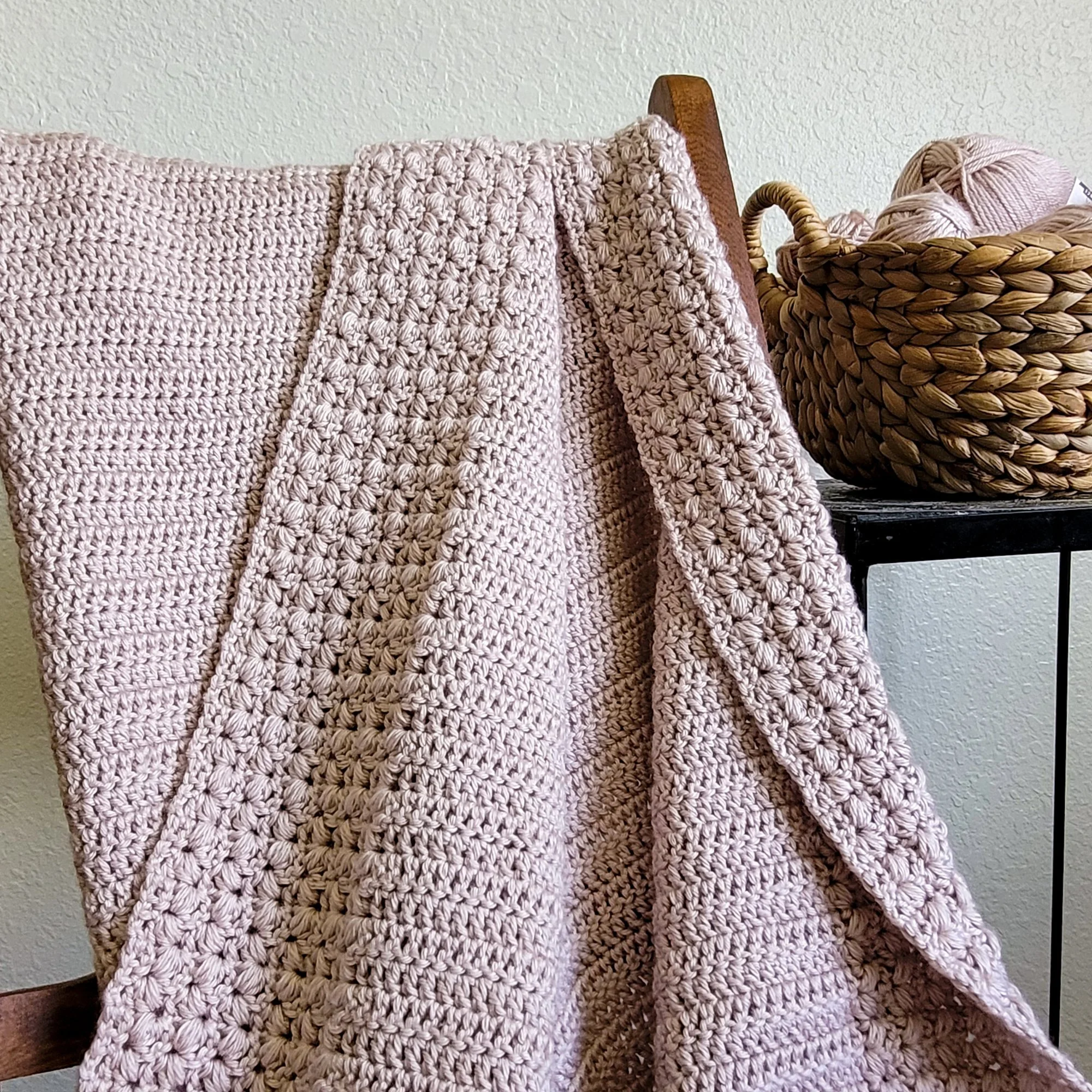 A pink textured crochet blanket draped over the back of a wooden rocking chair. Behind the chair and off to the right sits a table with a woven basket filled with pink yarn balls.
