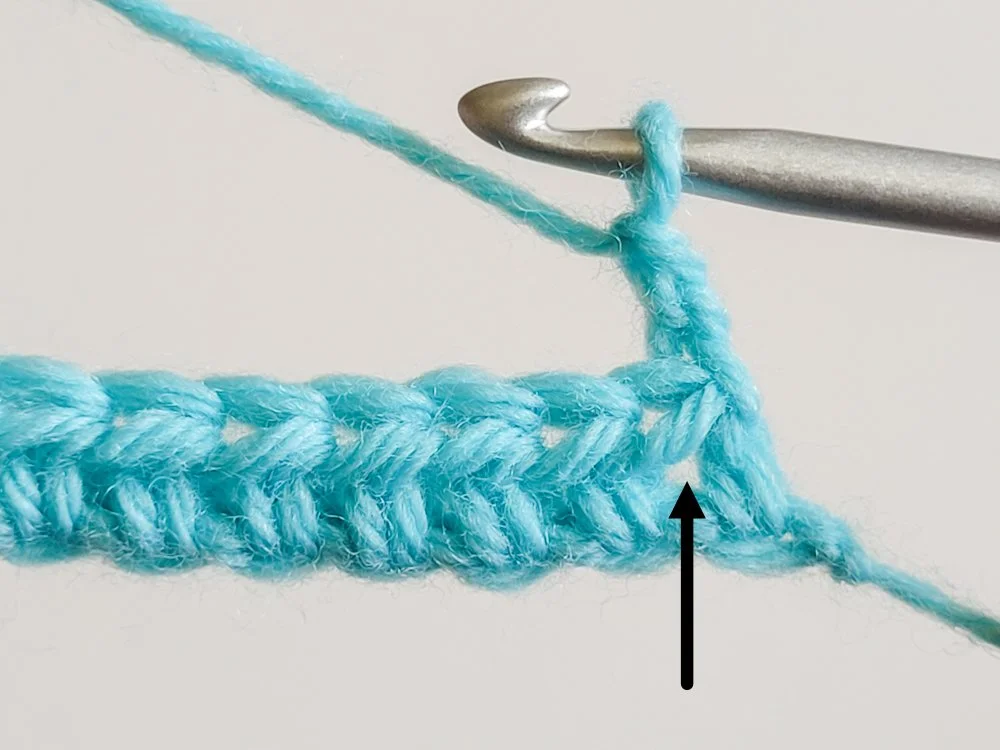 A chain 2 has been made to start row 2. A black arrow points to the space between the first and second stitch, where the first hdc will be worked.