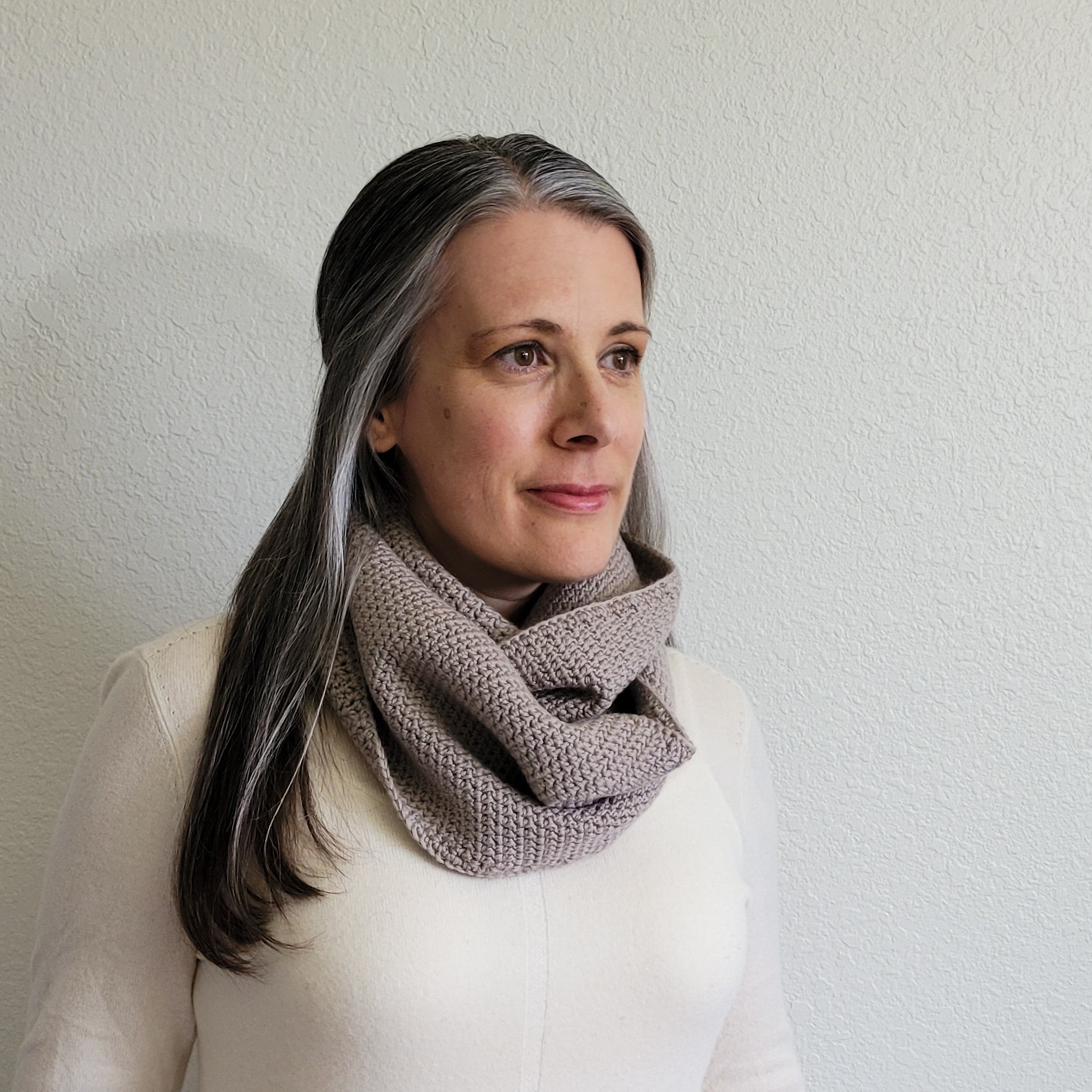 A woman with long, dark gray hair in a white sweater wears a grayish beige shawl wrapped casually several times around the neck.