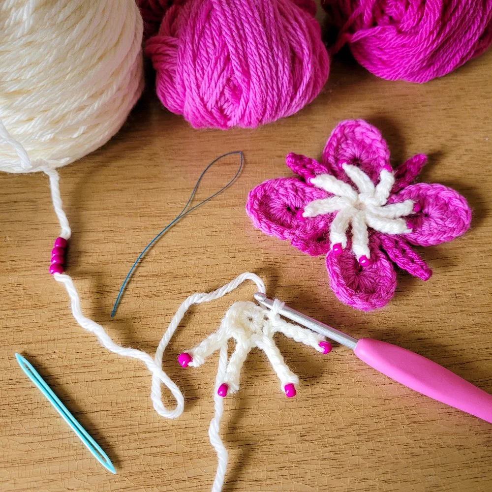 A completed crochet fireweed wildflower alongside one in progress. The completed flower is made with white and pink yarns. Magenta colored seed beads sit at the end of each chain in white to represent the flower's anthers.