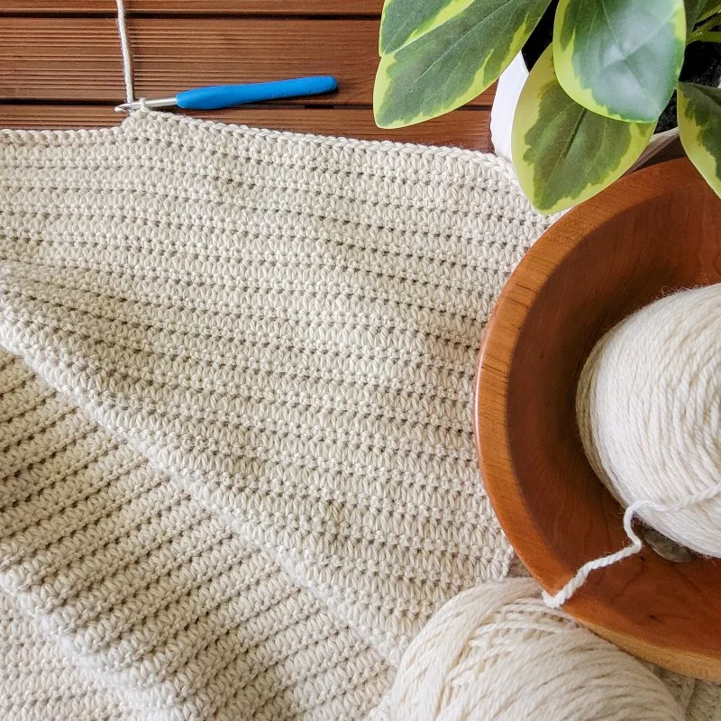 A crochet blanket in progress. The Forked Cluster crochet stitch creates a ribbed texture across the fabric. A yarn ball in a wooden bowl, a crochet hook, and a plant sit to the right of the blanket..