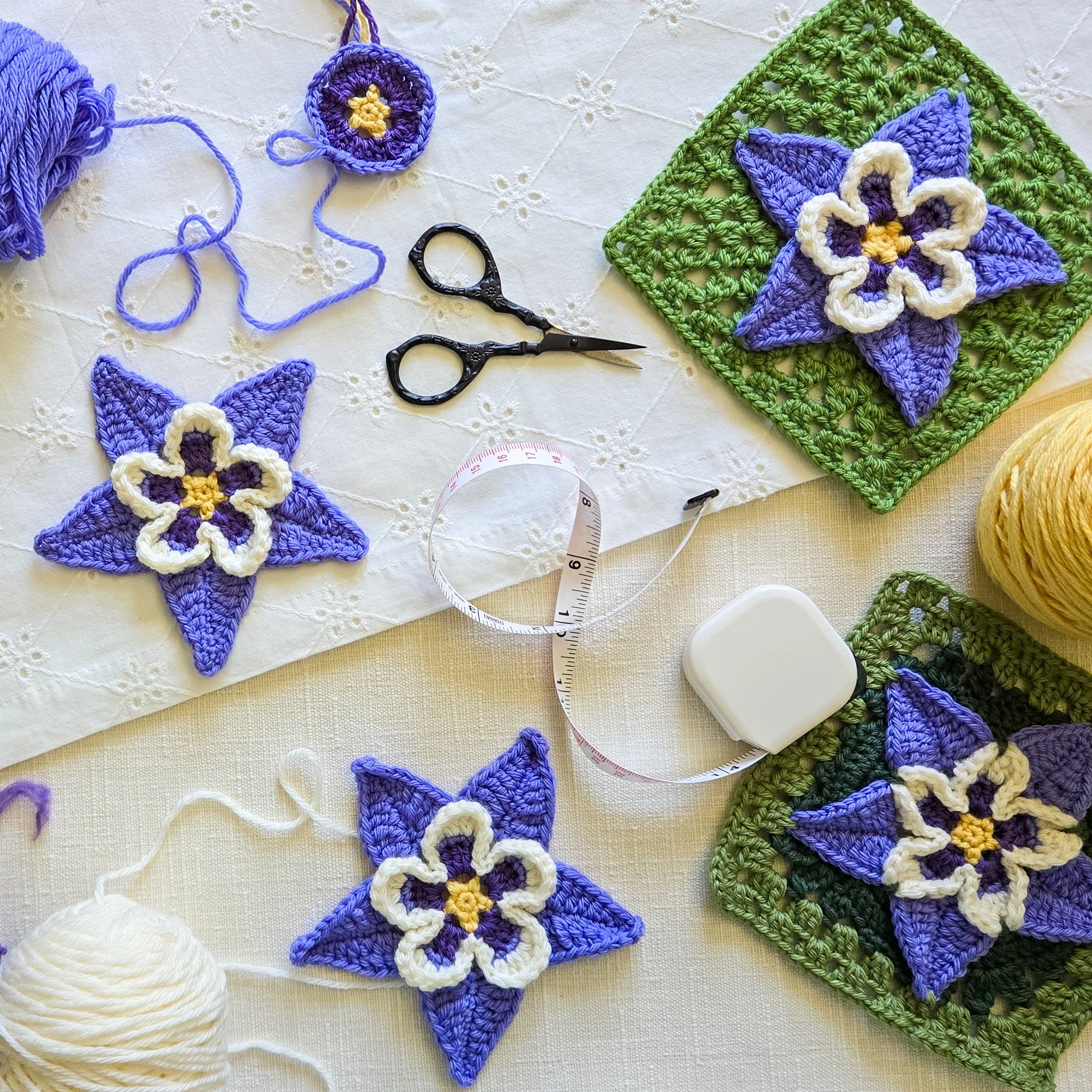 Several crochet columbine appliques and motifs with green crochet backgrounds are displayed on an eyelet tablecloth. Balls of yarn, a crochet hook, and other tools are also displayed.