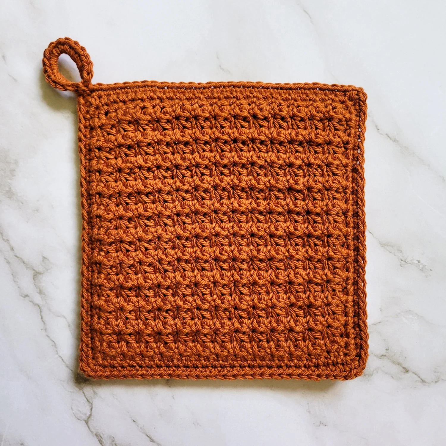Rust-colored Classic Potholder crocheted in dense, double-thick Trinity Stitch fabric with a hanging loop. The potholder is displayed on a white marble countertop.
