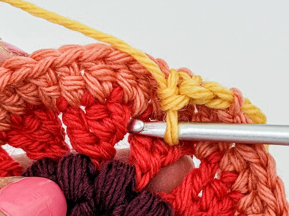 The outer edge of a crochet circle made with coral-colored yarn. A few single crochet have been worked in yellow. The hook is inserted into the space between red petals 2 rounds below.