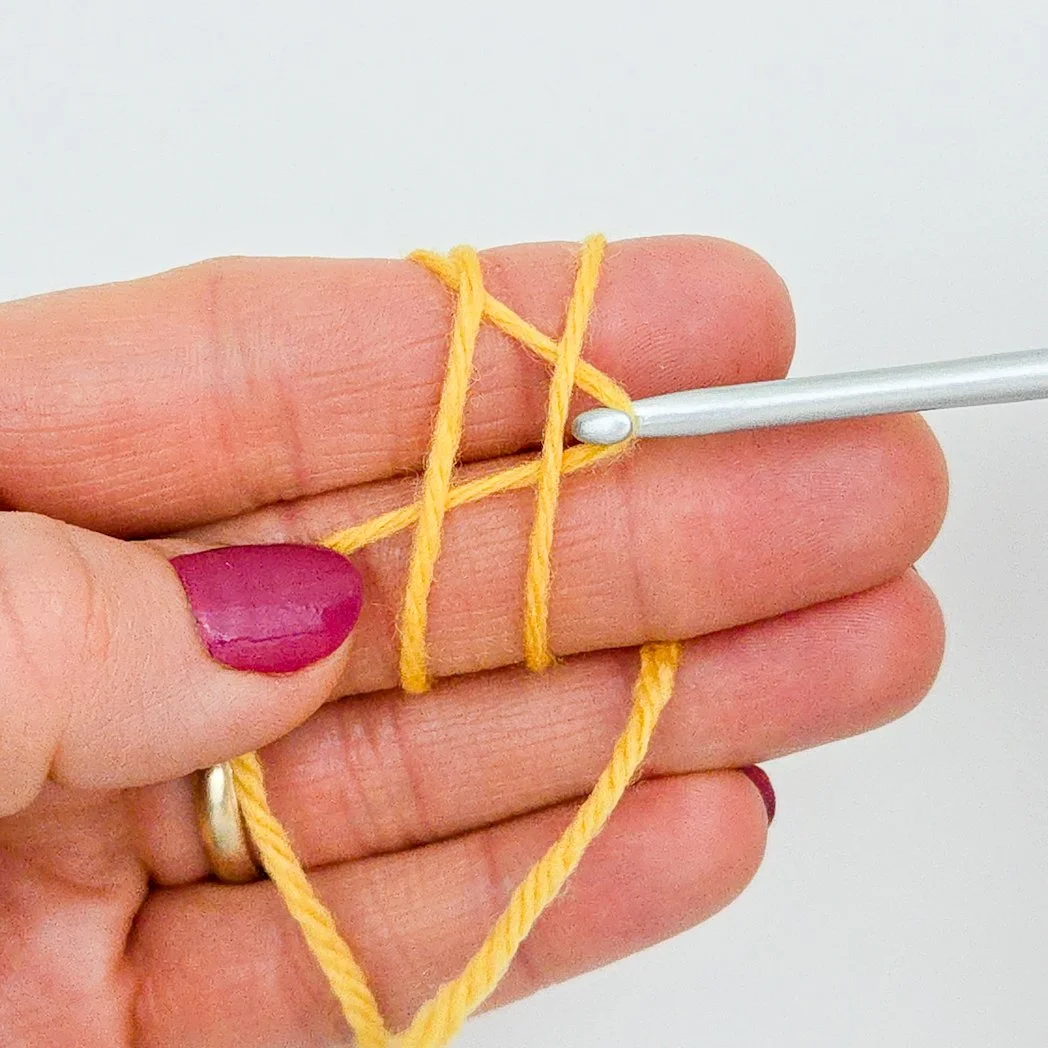 A strand of yellow yarn wrapped around two fingers. A crochet hook grabs the third strand and pulls it under the other two.