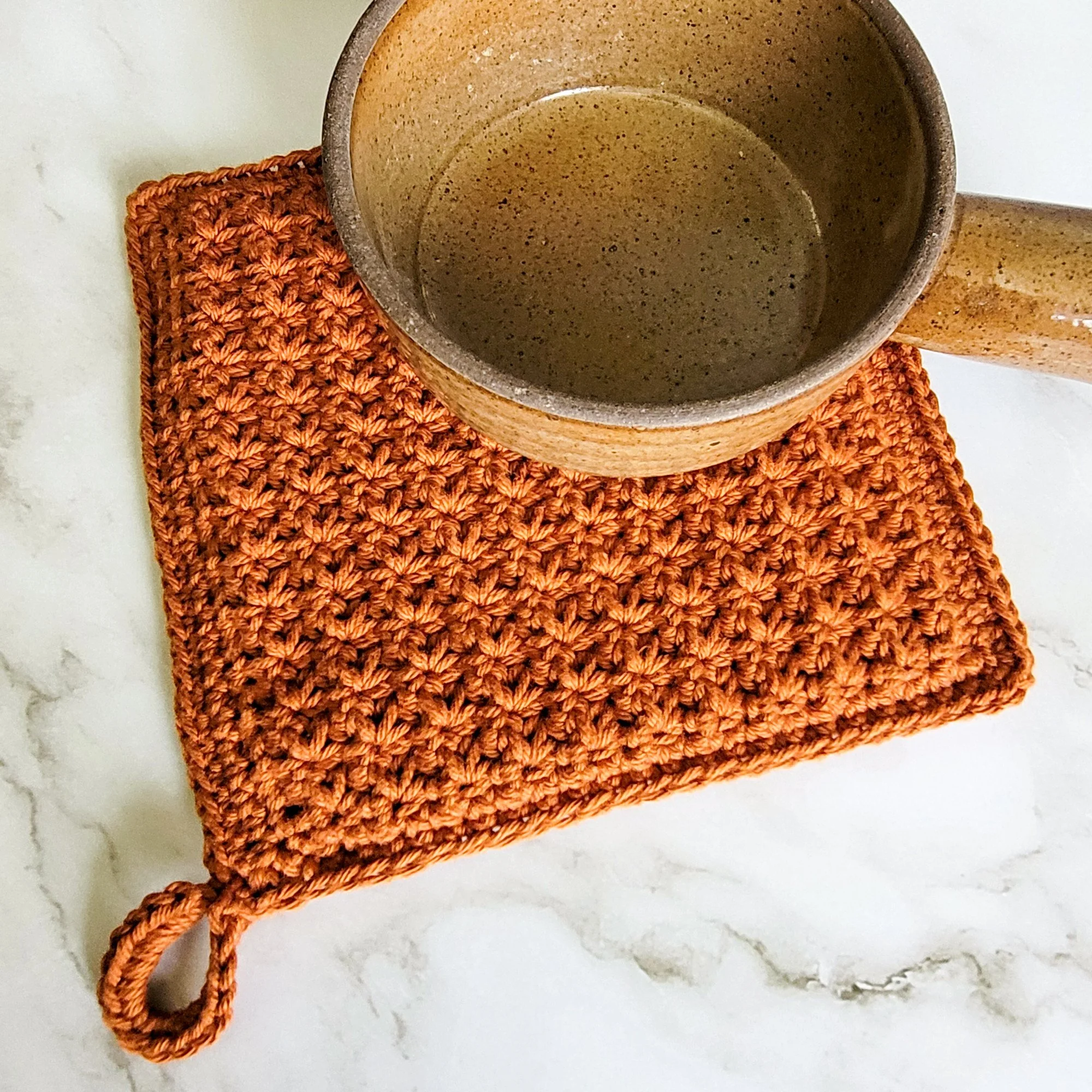 Rust-colored Classic Potholder crocheted in dense, double-thick Trinity Stitch fabric with a hanging loop. A clay bowl rests on top of the potholder, which is displayed on a white marble countertop.