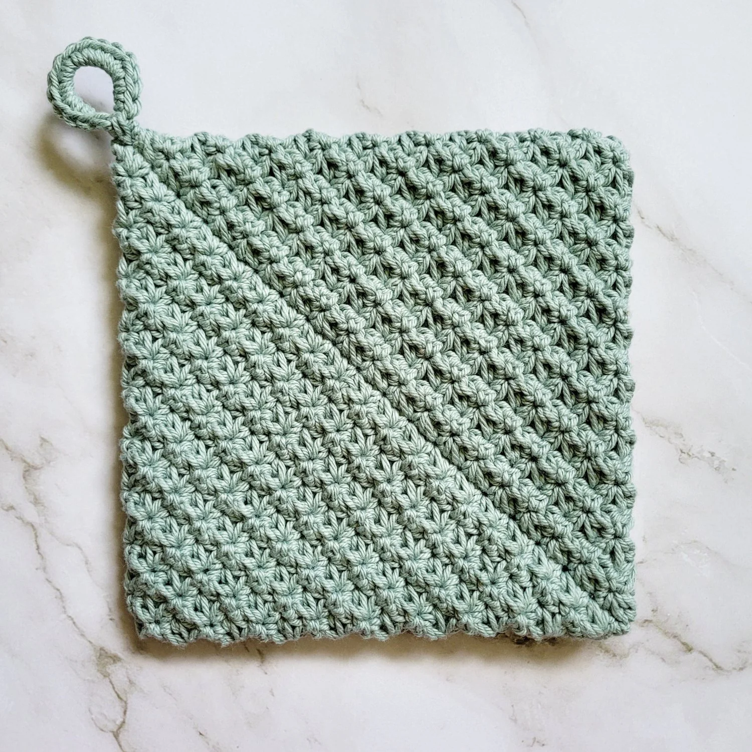 Sage green Folded Potholder crocheted in dense, double-thick Trinity Stitch fabric with a hanging loop. The folded construction creates a distinctive diagonal seam across the front. The potholder is displayed on a white marble countertop.