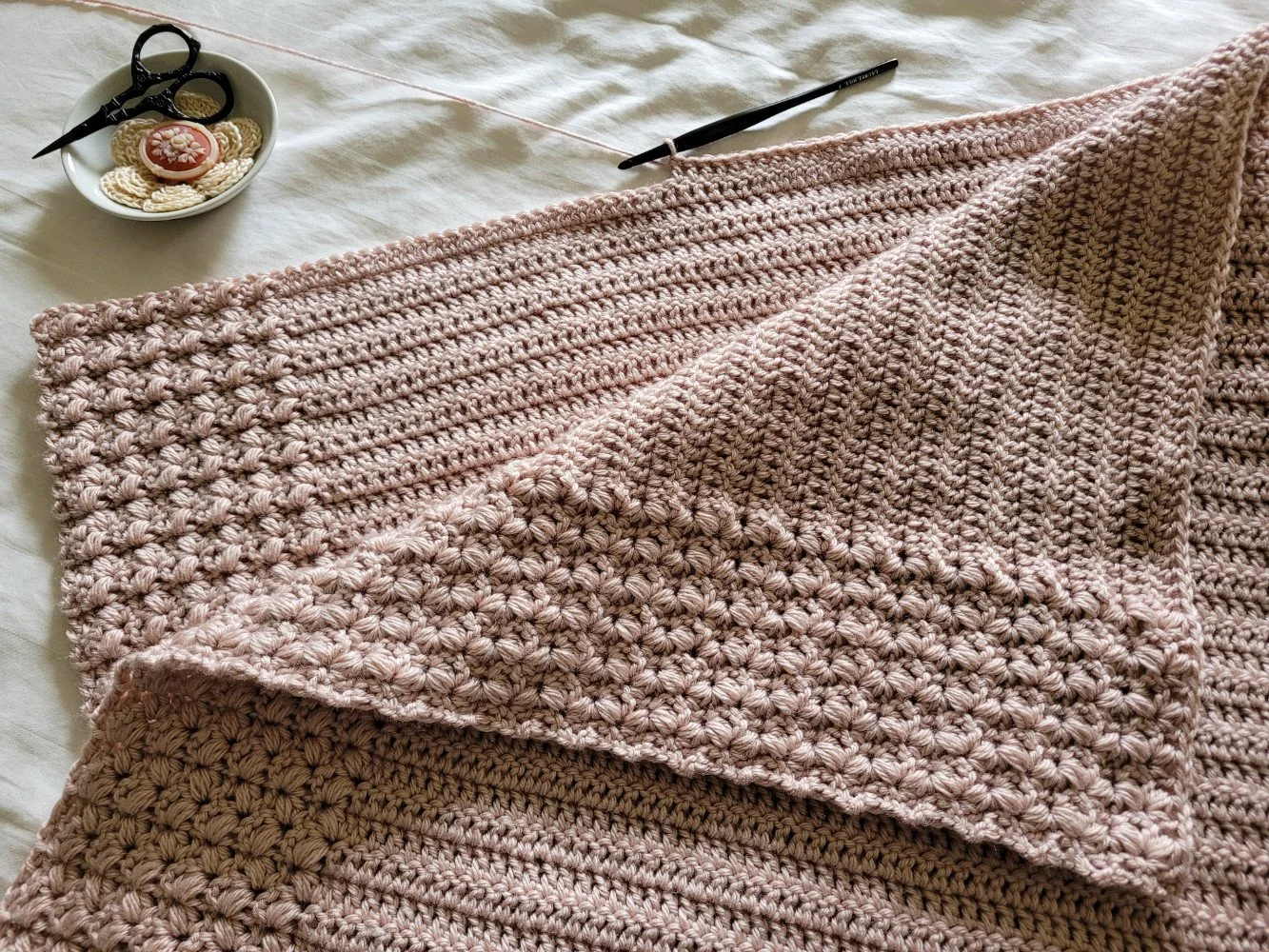 A pink textured crochet blanket in progress. The border features soft puff stitches, while the center panel is made with sleek double crochet.