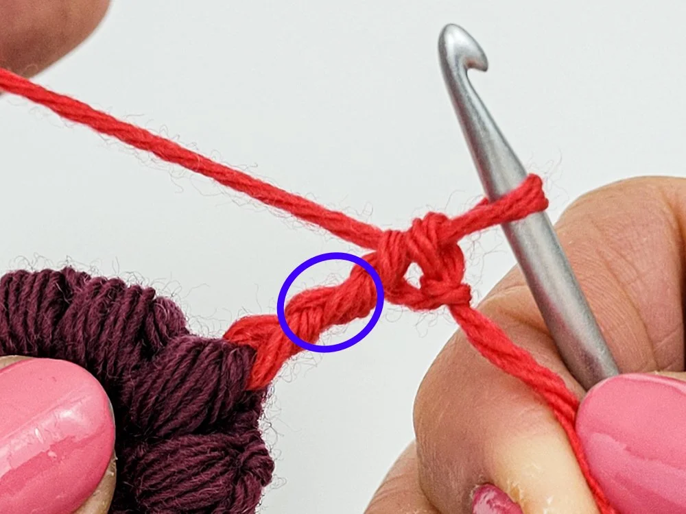 A red yarn is attached to a purple crochet circle with a standing treble crochet. A blue circle has been drawn around the center of the stitch.
