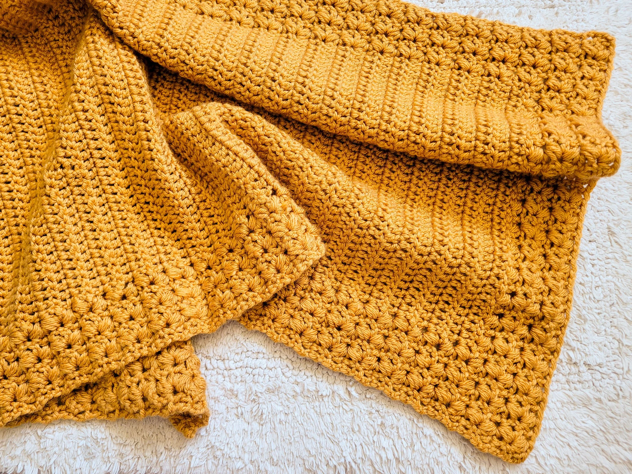 A mustard-yellow crochet blanket featuring a textured border made with puff V-stitches. The blanket is loosely arranged across a white rug to show off the soft drape of the fabric.