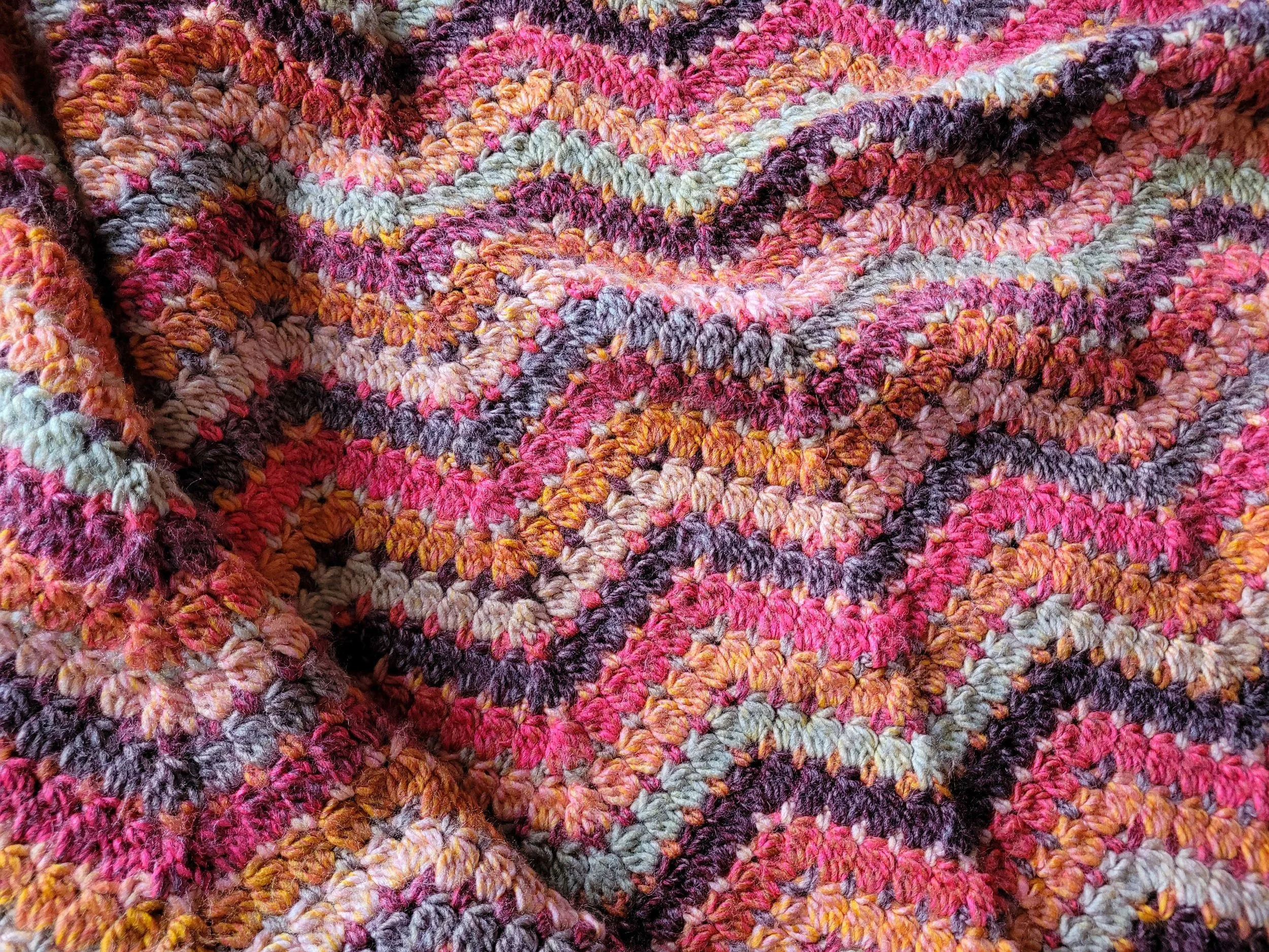 Close-up of a crochet ripple blanket with wavy stripes in shades of red, orange, brown, and pale green, with the fabric folded in the bottom-left corner.