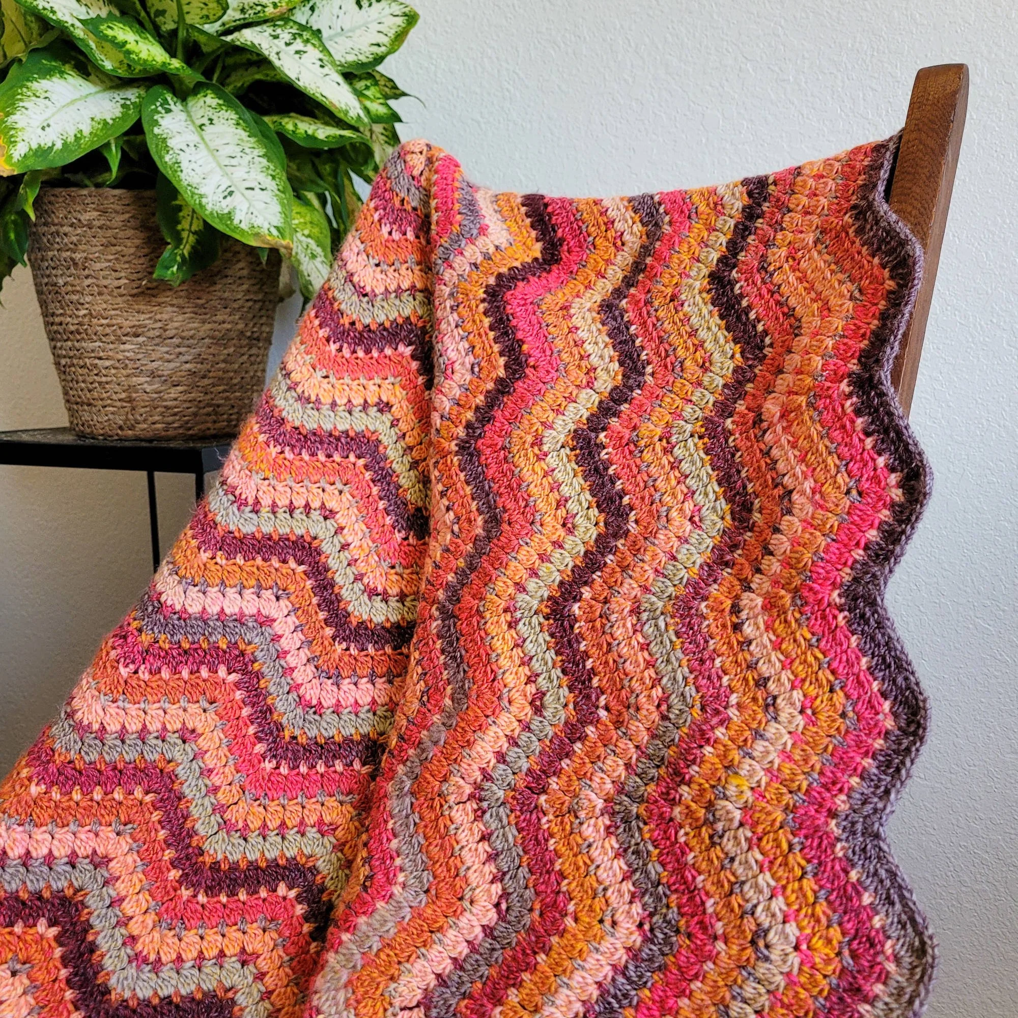 A crochet ripple blanket with wavy stripes in shades of red, orange, brown, and pale green is draped over a chair. A leafy green plant on a metal stand sits behind the blanket to the left.