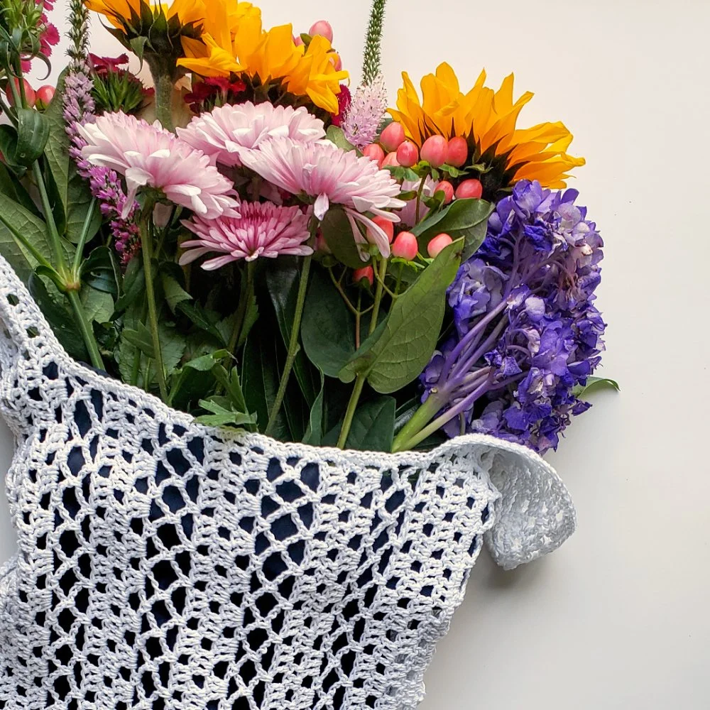 A white crochet lace market bag filled with a colorful bouquet of  fresh spring flowers.