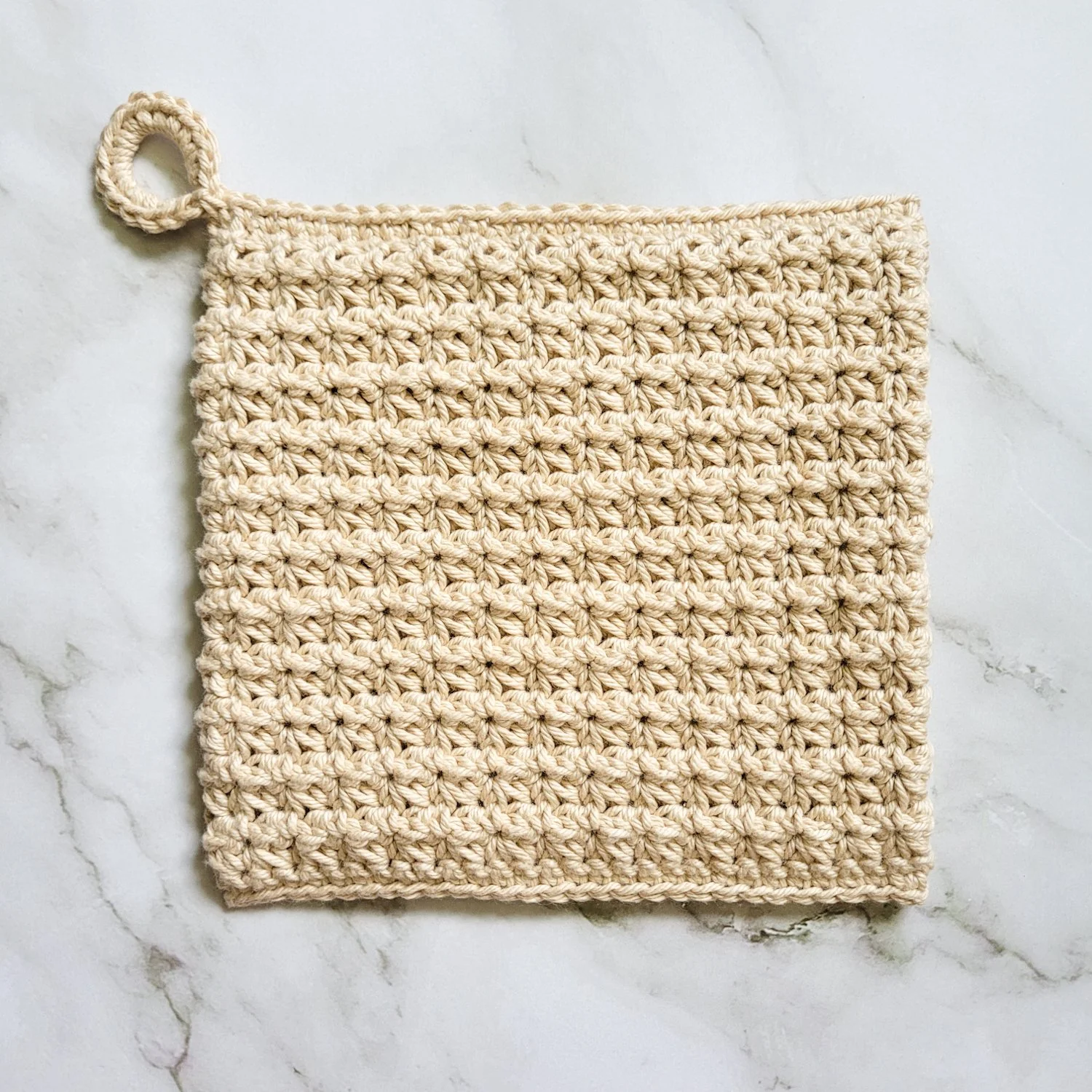 Beige Ring-Start Potholder crocheted in dense, double-thick Trinity Stitch fabric with a hanging loop. The potholder is displayed on a white marble countertop.