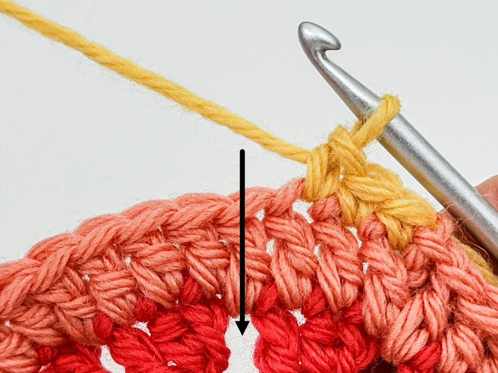 The outer edge of a crochet circle made with coral-colored yarn. A few single crochet have been worked in yellow. A black arrow points to the space between red petals 2 rounds below, indicating where to insert hook next.