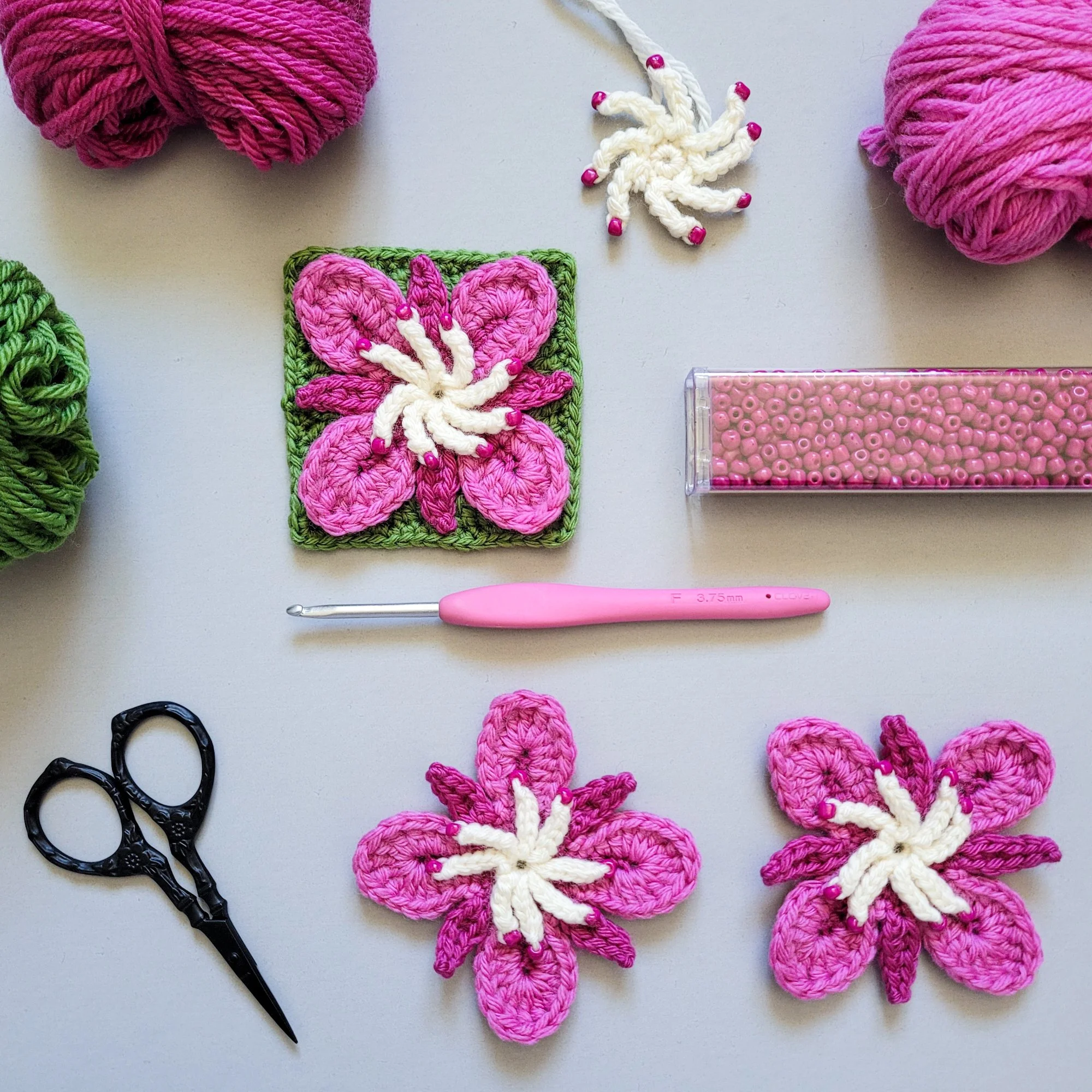 Several bright pink crochet flowers (alongside balls of yarn, a crochet hook with a pink handle, and a tube of magenta colored beads) displayed on a gray background.