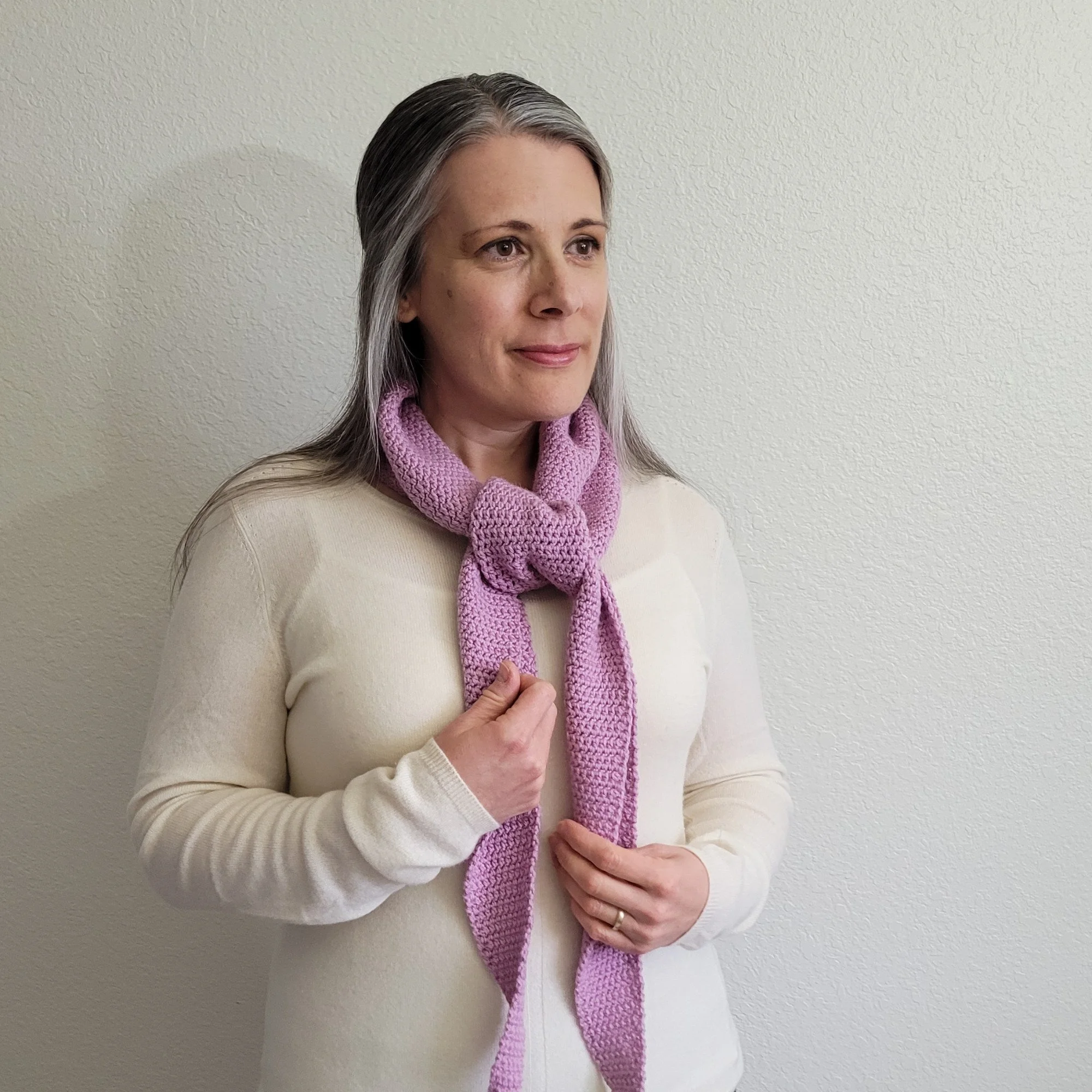 A woman with long, gray hair in a white sweater wears a light purple scarf tied in an overhand knot.