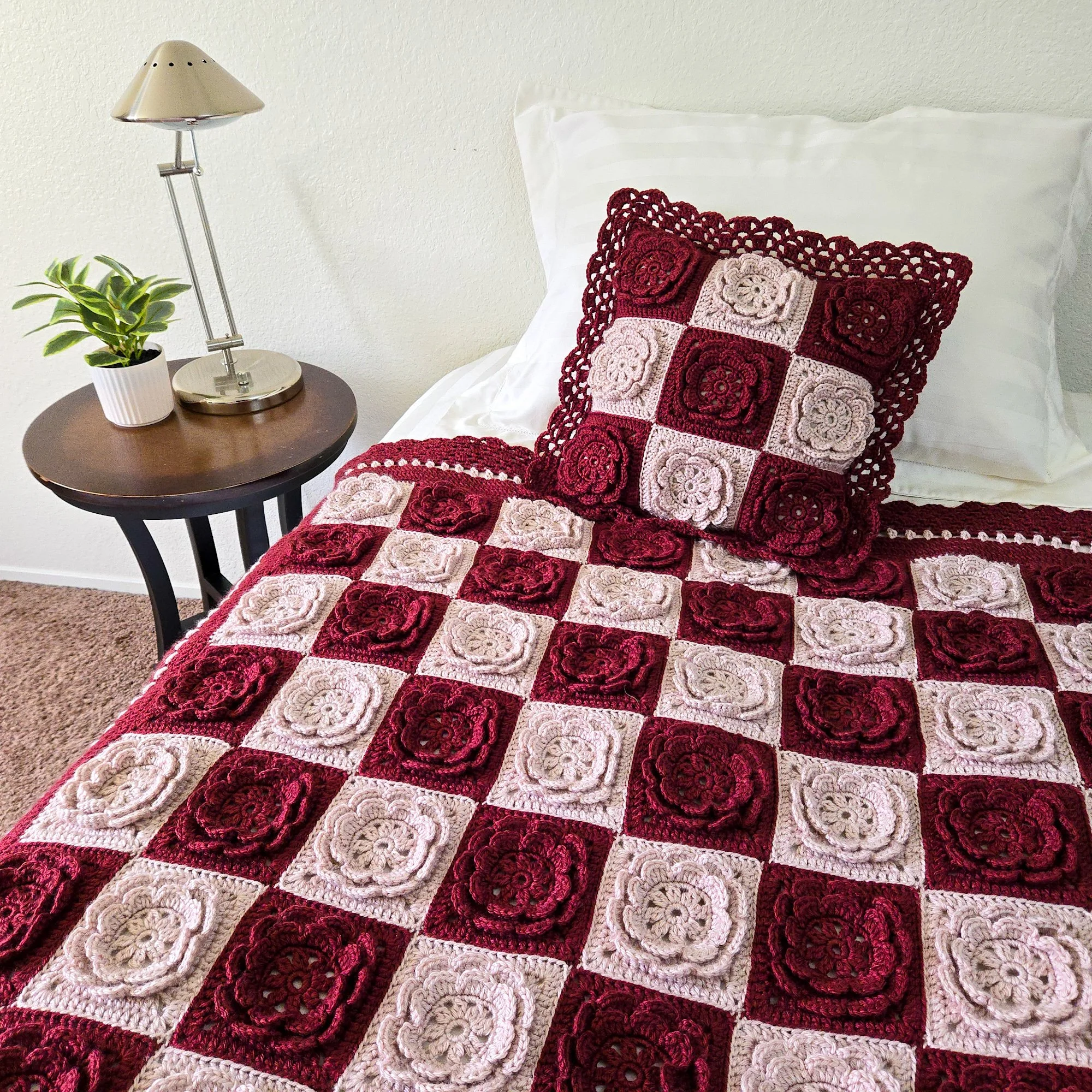 Crochet throw blanket made with square rose motifs in deep red and blush pink arranged in a striking checkerboard pattern is draped across a bed with white linens. Blanket is edged in a lacy border stitched in red and pink. A matching pillow featurin
