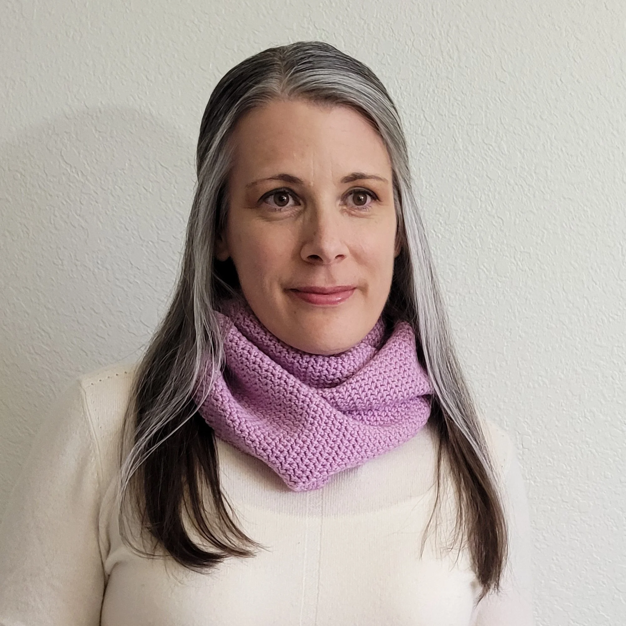 A woman with long, gray hair in a white sweater wears a light purple scarf wrapped casually several times around the neck.