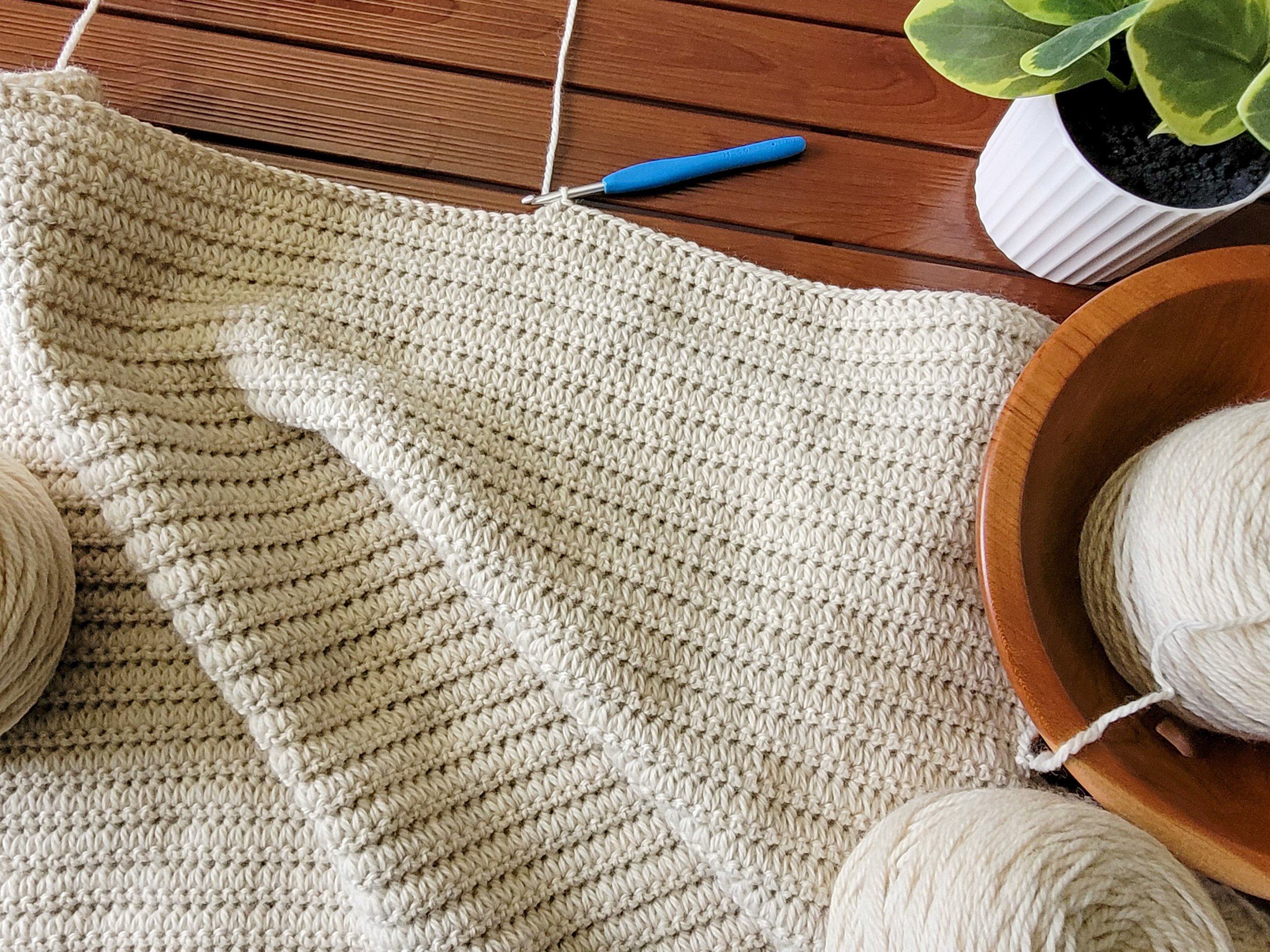 In-progress photo of the Hand in Hand Blanket. The forked cluster crochet stitch creates a ribbed texture across the fabric. A yarn ball in a wooden bowl, a crochet hook, and a plant sit to the right of the blanket.