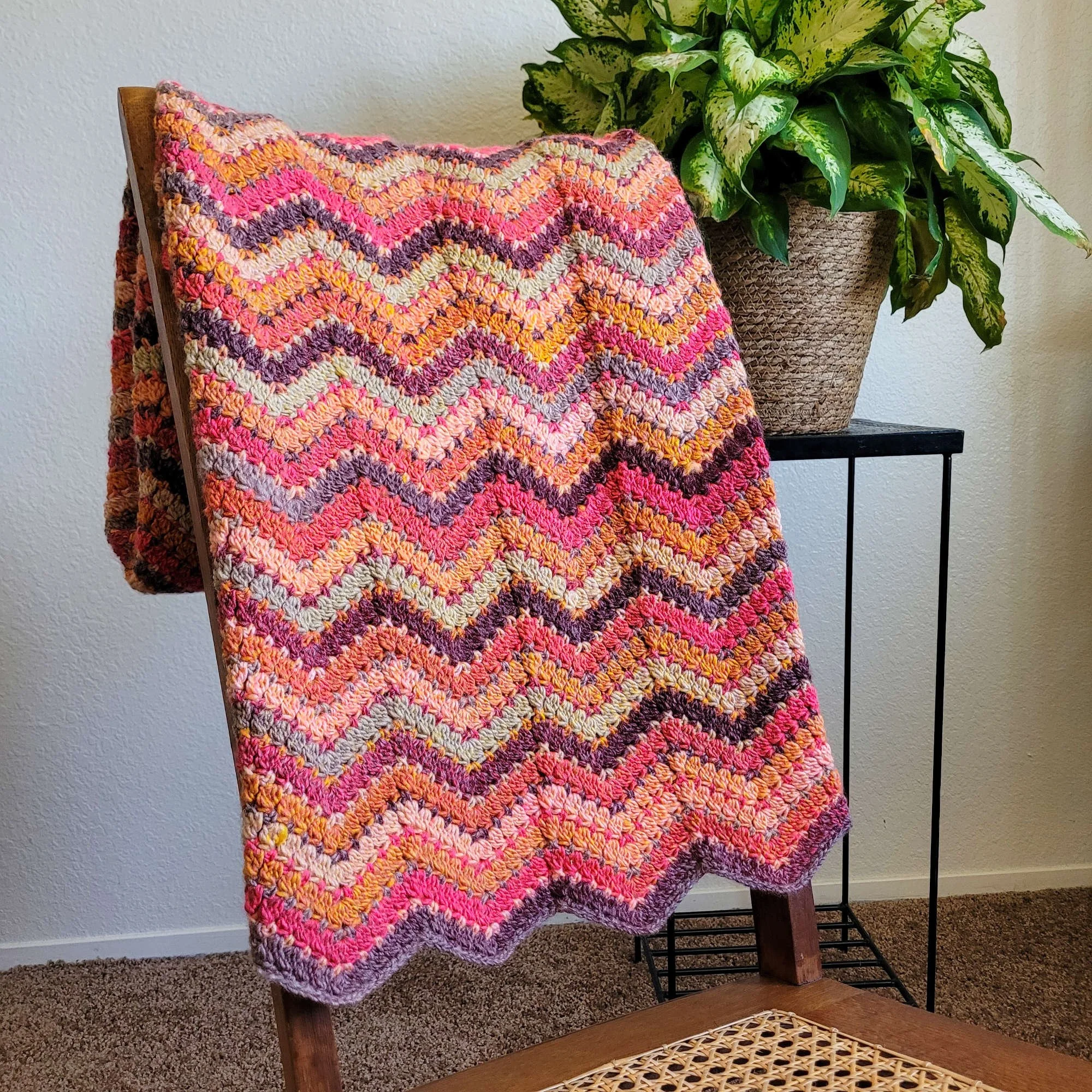 A crochet ripple blanket with wavy stripes in shades of red, orange, brown, and pale green is folded and draped neatly over the back of a rocking chair. A leafy green plant on a metal stand sits behind the blanket to the right.