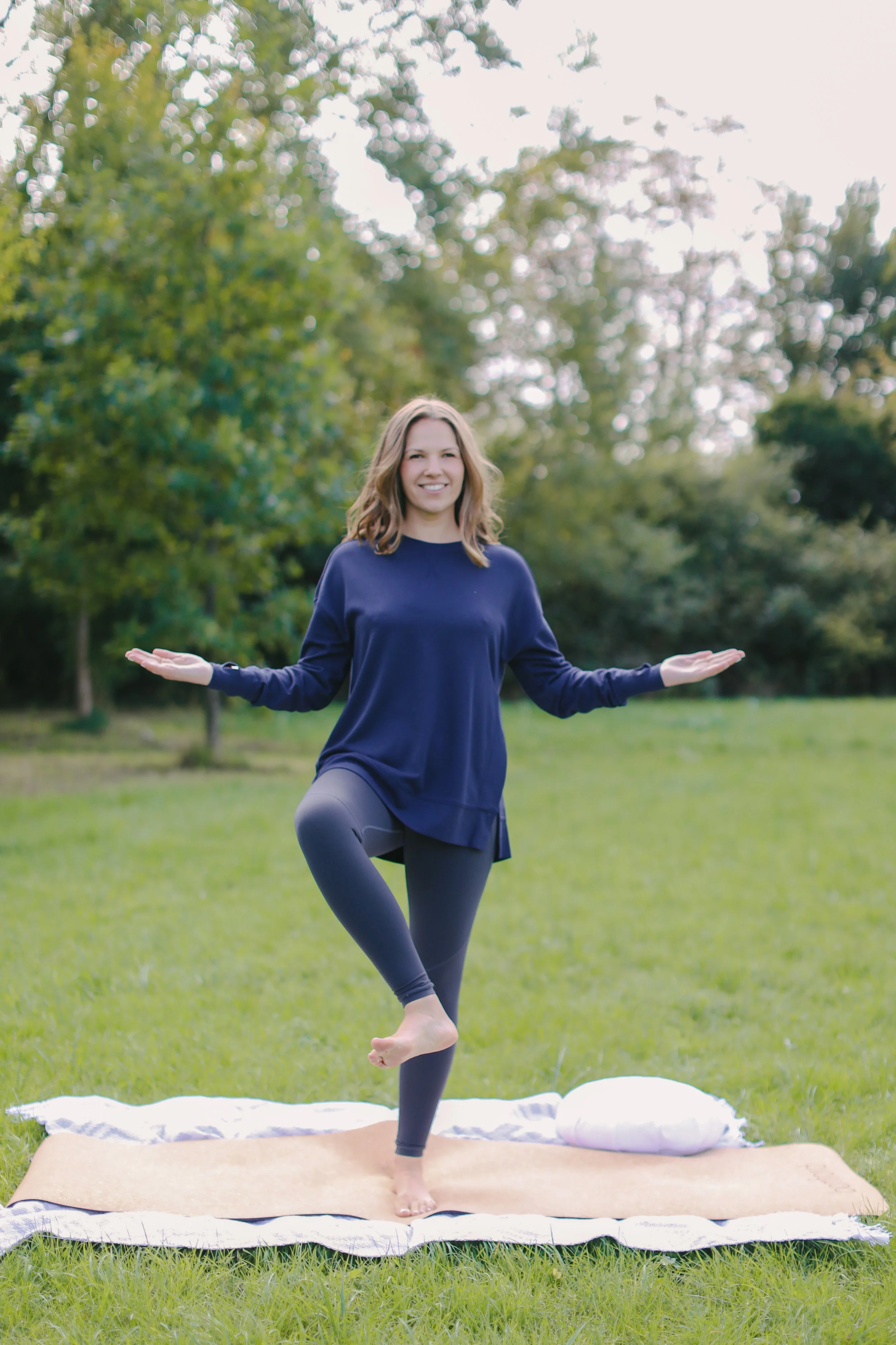 A person practicing a yoga pose outdoors on a yoga mat, standing on one leg with arms extended. They are wearing a blue top and leggings, surrounded by grass and trees.