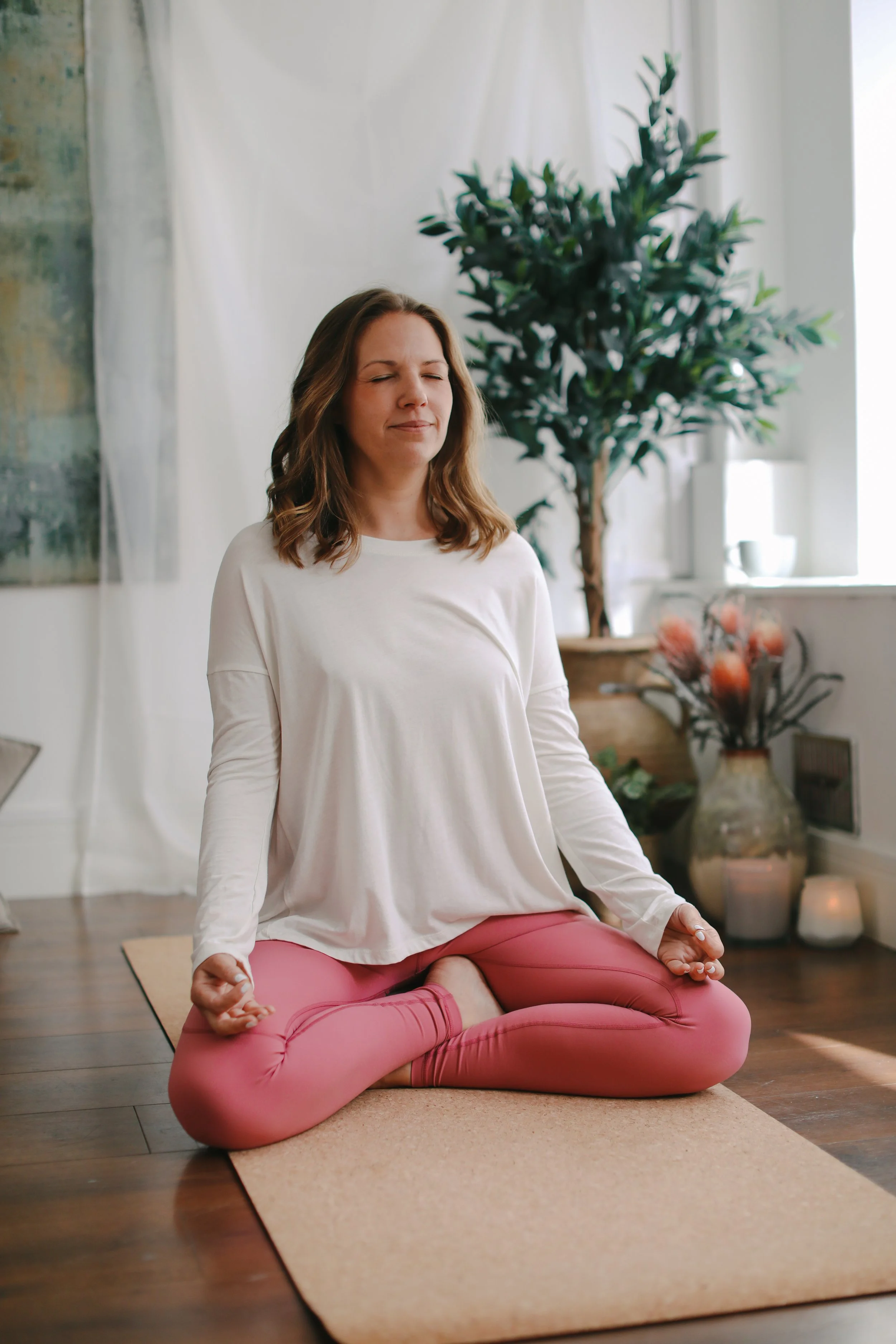 Woman meditating in a seated yoga pose on a mat indoors, wearing a white shirt and pink leggings, with eyes closed, near a plant and soft lighting.
