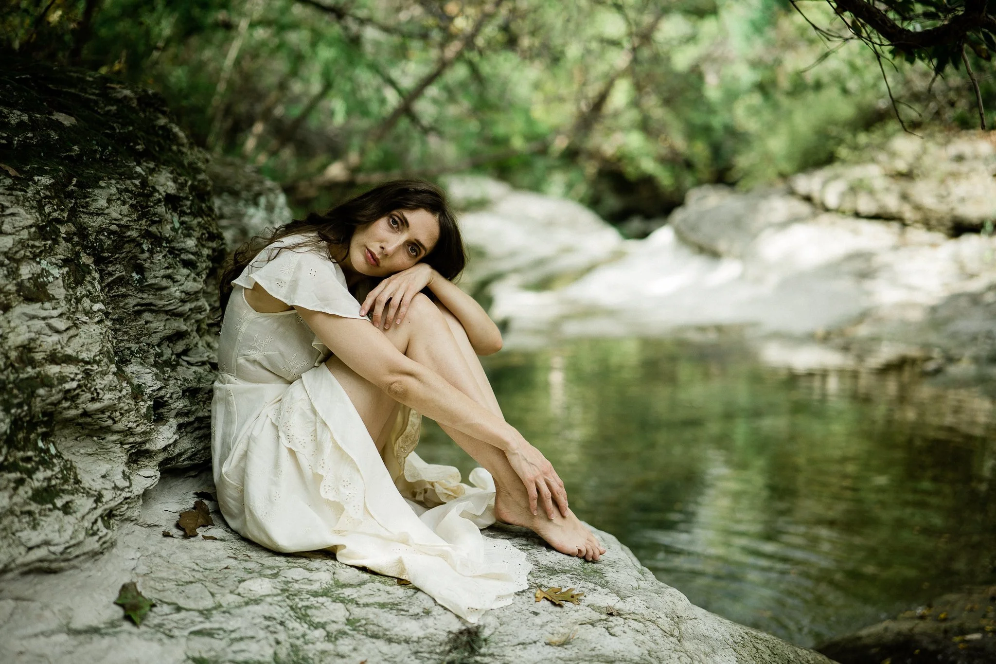 Portrait of a female with brown hair wearing a cream colored dress. She is sitting down on rocks with water behind her