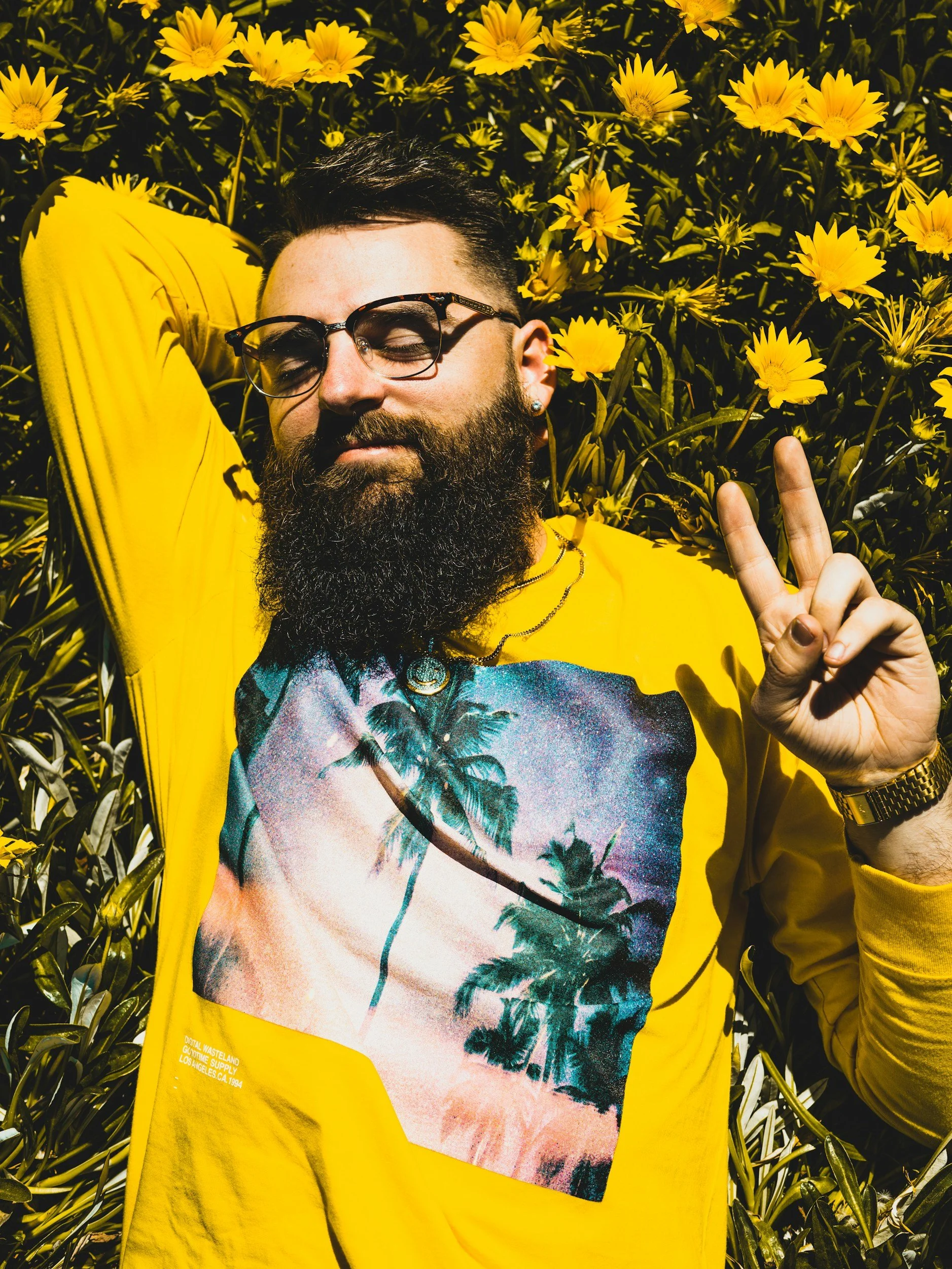 A man with dark hair, glasses, and a beard, wearing a yellow shirt and a graphic T-shirt with palm trees and a sunset, makes a peace sign with his right hand while lying among yellow flowers with green leaves.