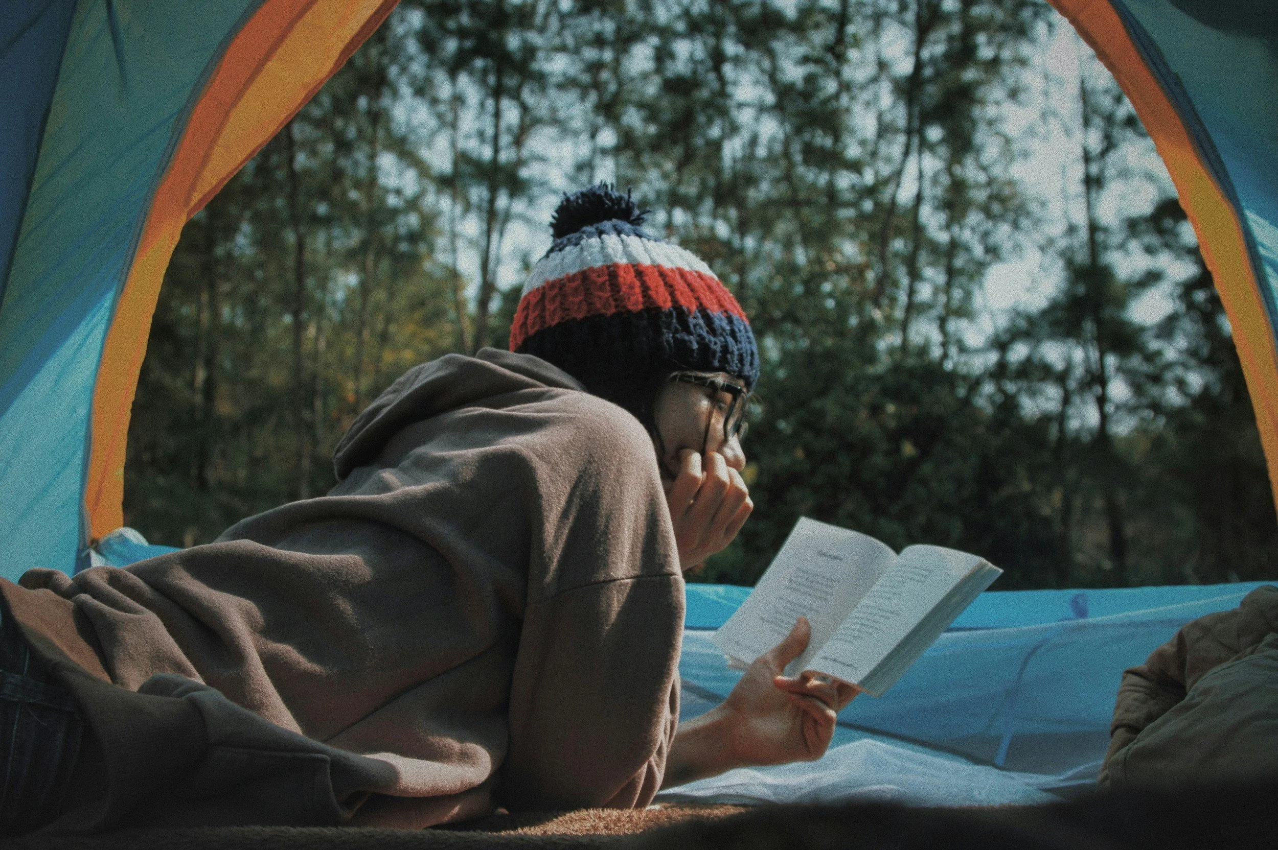 Person wearing a multicolored knit hat and glasses, sitting inside a tent outdoors, reading a book.