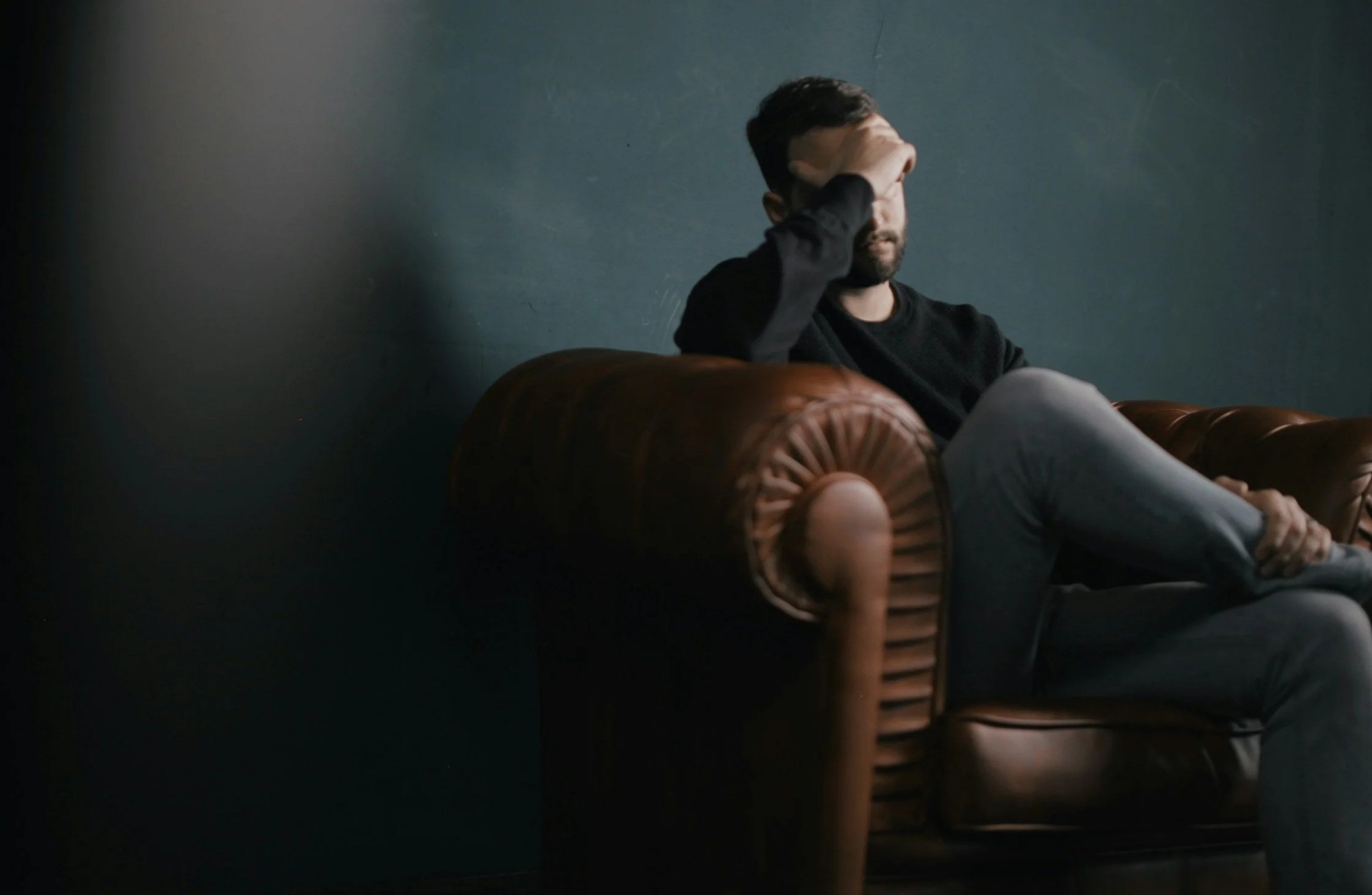 A man with depression sitting on a vintage brown leather couch against a dark blue wall, with his hand covering his face in a contemplative pose, wearing a black long sleeve shirt and gray pants.