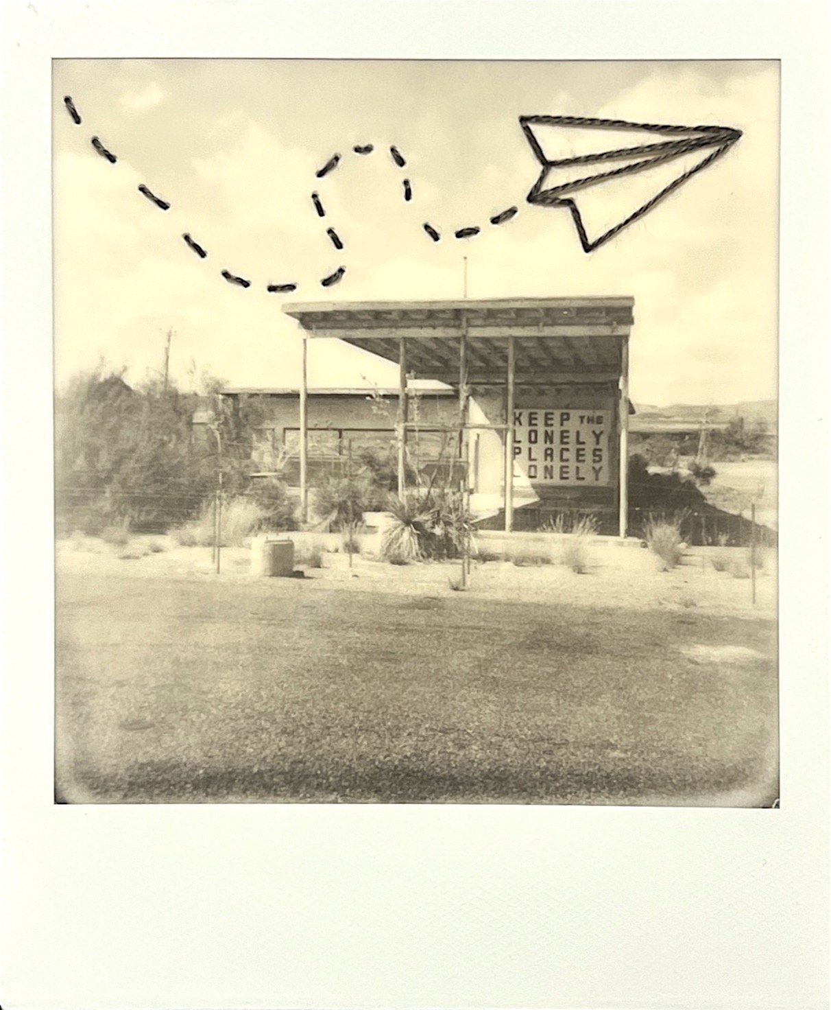 A Polaroid photograph of a building with a sign that reads 'KEEP THE LONELY PLACES LONELY,' taken outdoors on a partly cloudy day with desert-like surroundings.