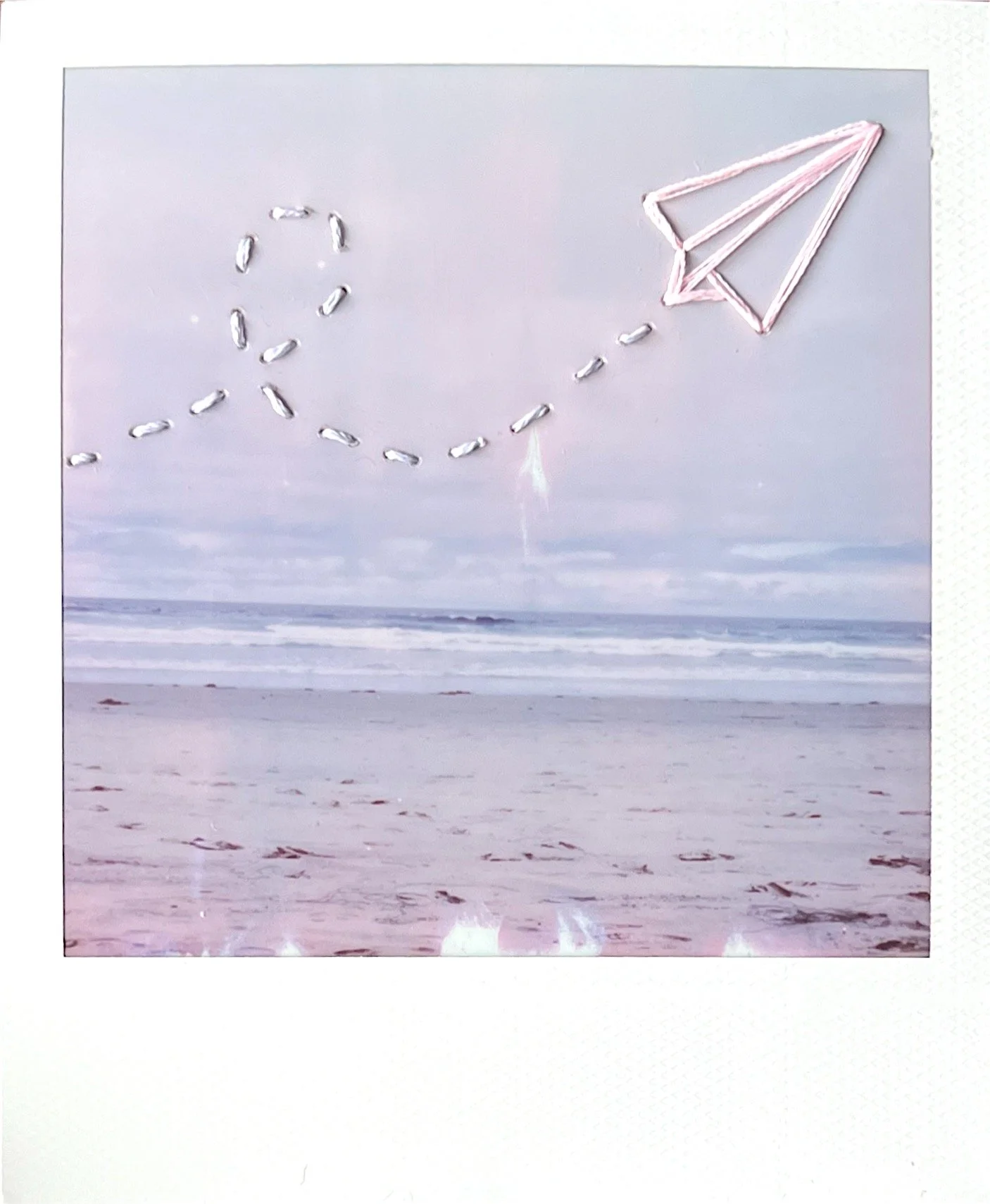 A photograph of ocean waves with a distant landmass and a sky background. The image has pink embroidery of a paper airplane and a dotted line indicating flight path.