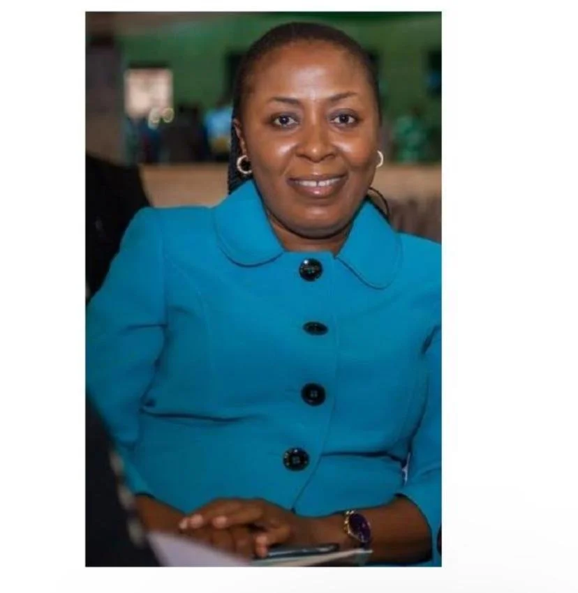 A middle-aged woman with short hair, wearing a bright blue blazer with black buttons and earrings, smiling and seated indoors.