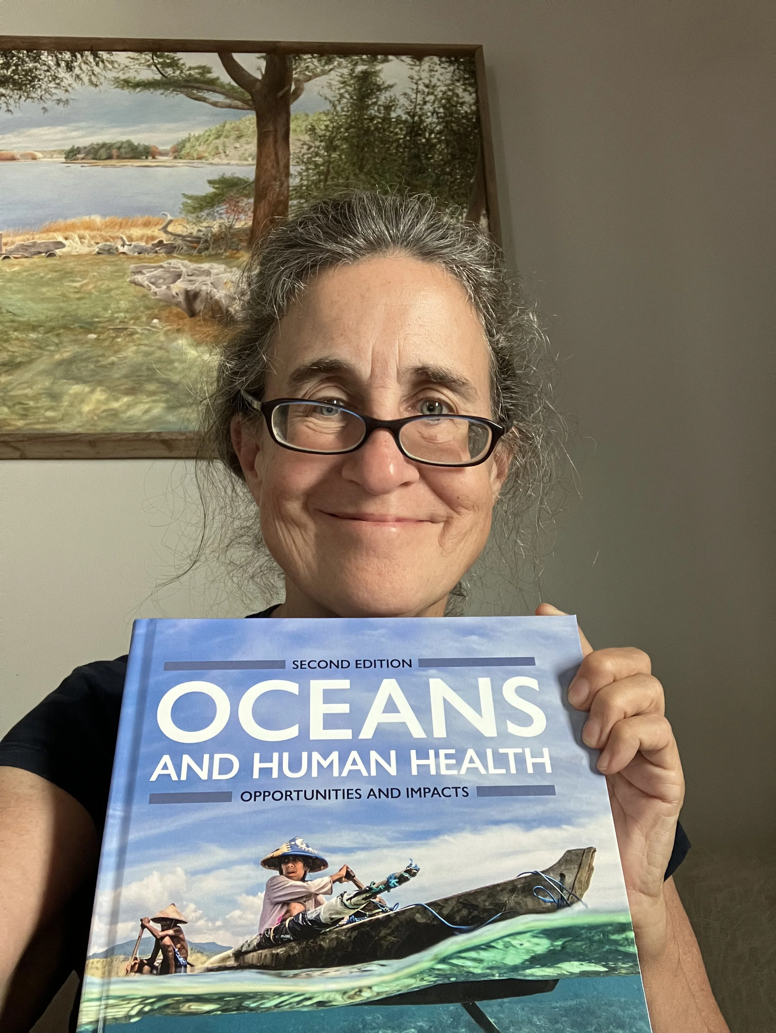 A woman with glasses and gray hair holding a book titled "Oceans and Human Health" in front of a gray wall with a landscape painting of a tree by the water