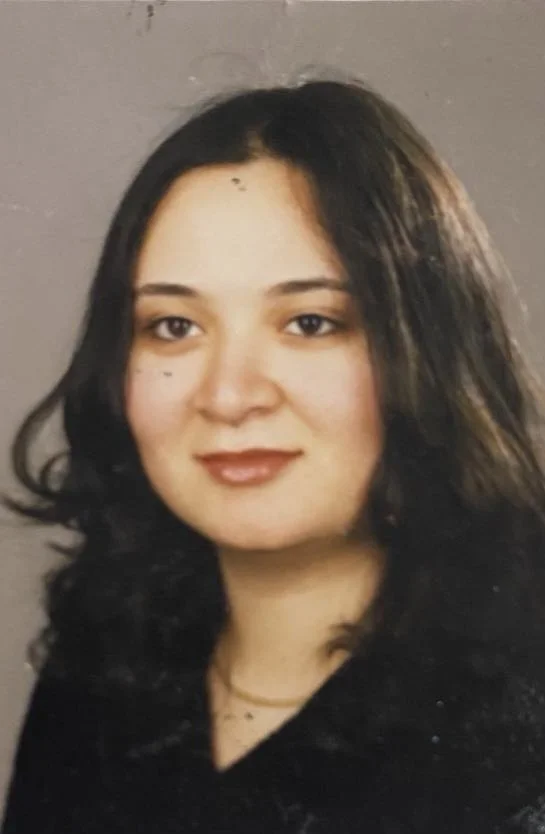 A young woman with shoulder-length dark hair, wearing a black top, and looking at the camera with a slight smile, against a plain grey background.