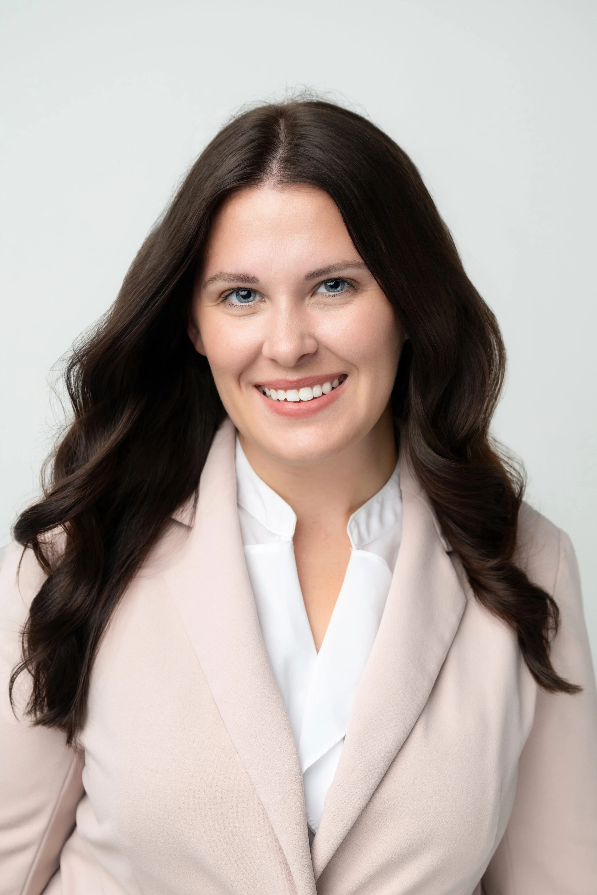 A woman with long dark hair, blue eyes, and a friendly smile, wearing a beige blazer over a white blouse, against a plain light background.
