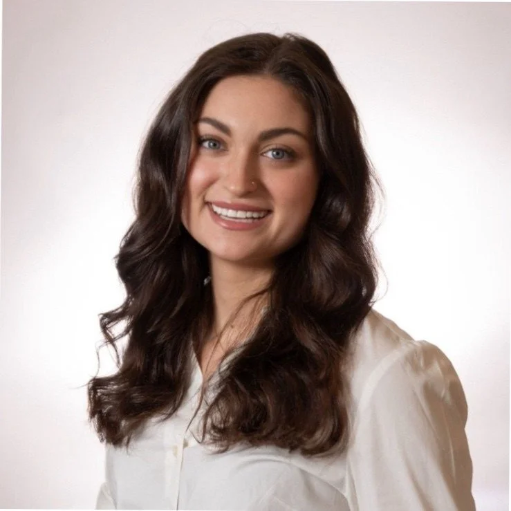 A young woman with long dark brown wavy hair, blue eyes, and a warm smile, wearing a white blouse against a plain white background.