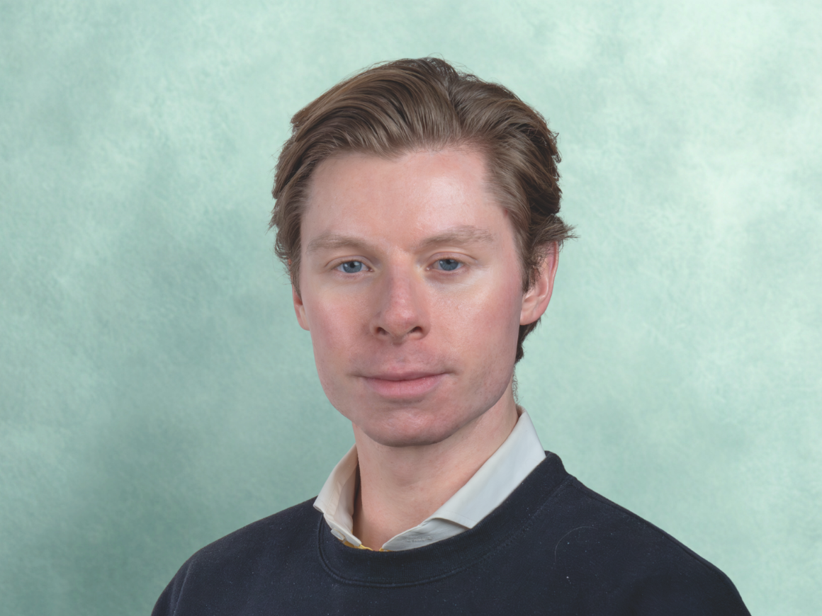 A young man (filmmaker Kyle Edwardson) with light skin, brown hair, and blue eyes wearing a black sweater over a collared shirt against a green background.