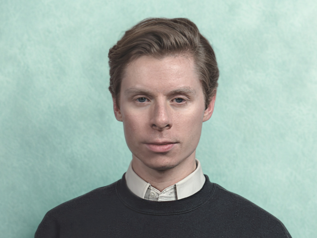 Portrait of a young man with light brown hair (Filmmaker Kyle Edwardson) wearing a black sweater over a white collared shirt against a soft green background.