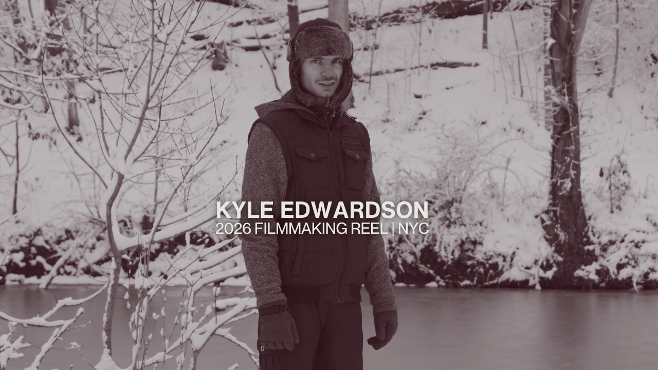 A man wearing winter clothing, including a fur-lined hat, standing outdoors in a snowy landscape near a body of water, with trees and snow-covered ground in the background.