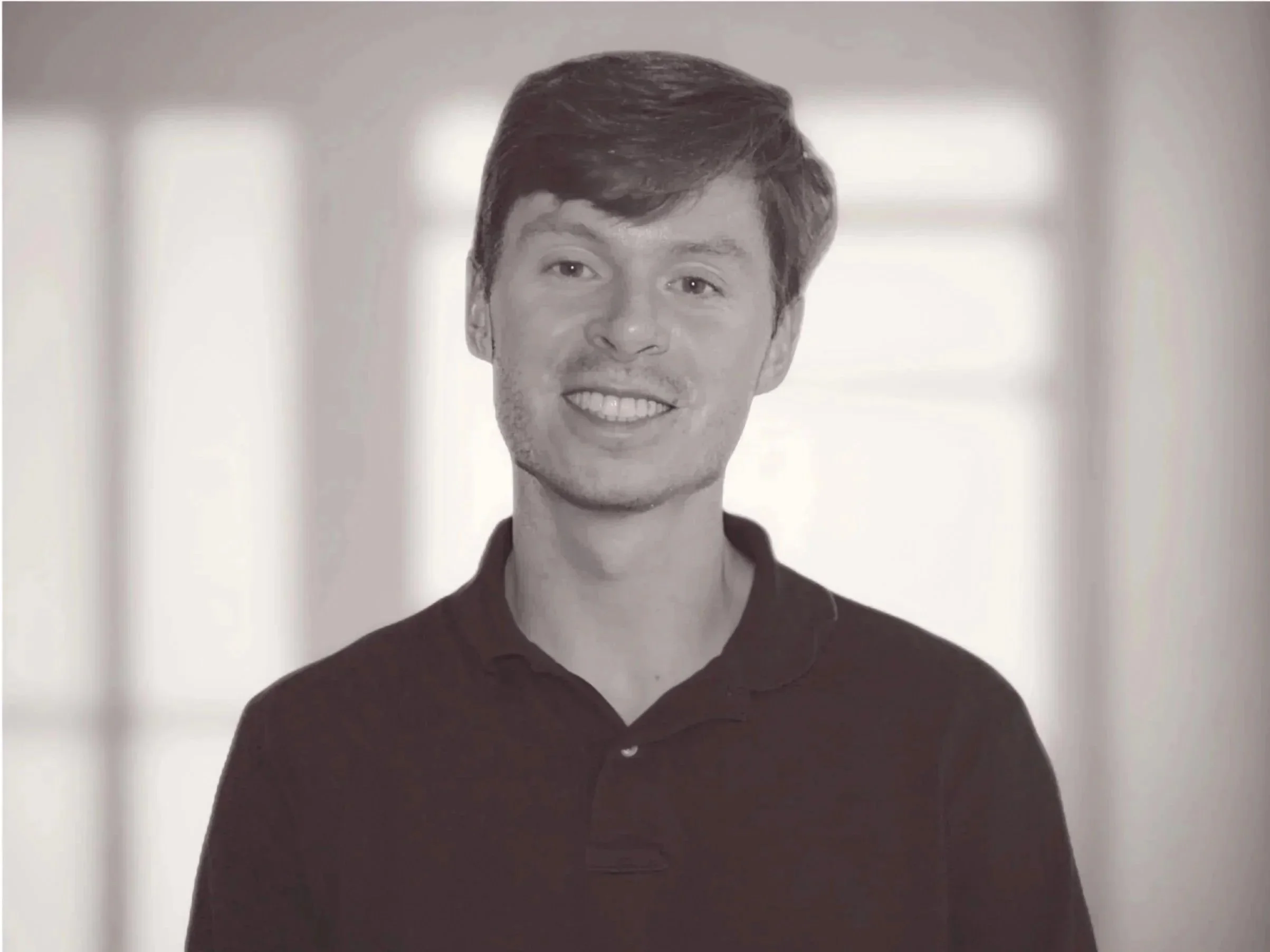 A black and white photograph of a young man with short, dark hair, smiling and wearing a collared shirt, standing in front of a blurred background with windows.