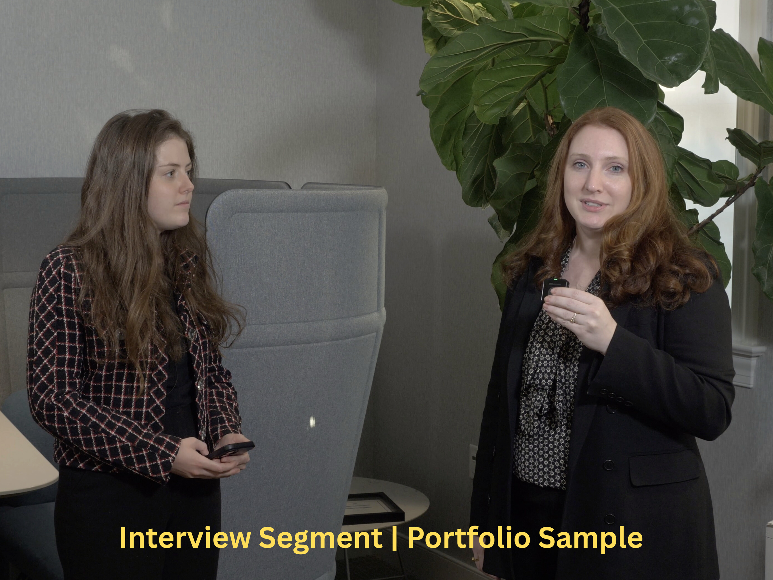 A woman is speaking, holding a microphone, while a young girl listens attentively, holding a smartphone, in an office setting with a large green plant in the background. Film production by Kyle Edwardson, owner of Hart Street Studios