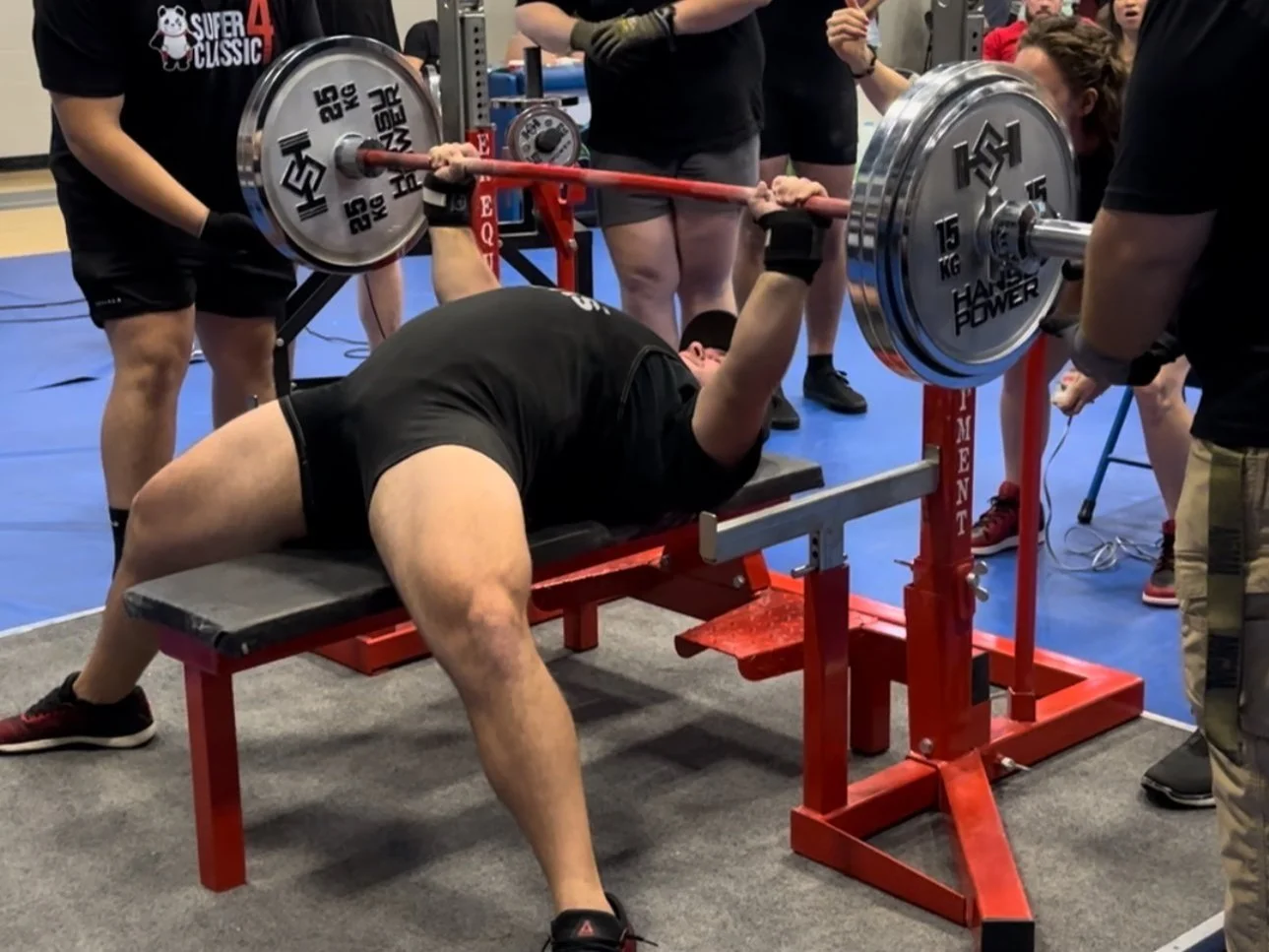 A man is performing a bench press with a barbell loaded with weights at a powerlifting competition, surrounded by spectators and officials.